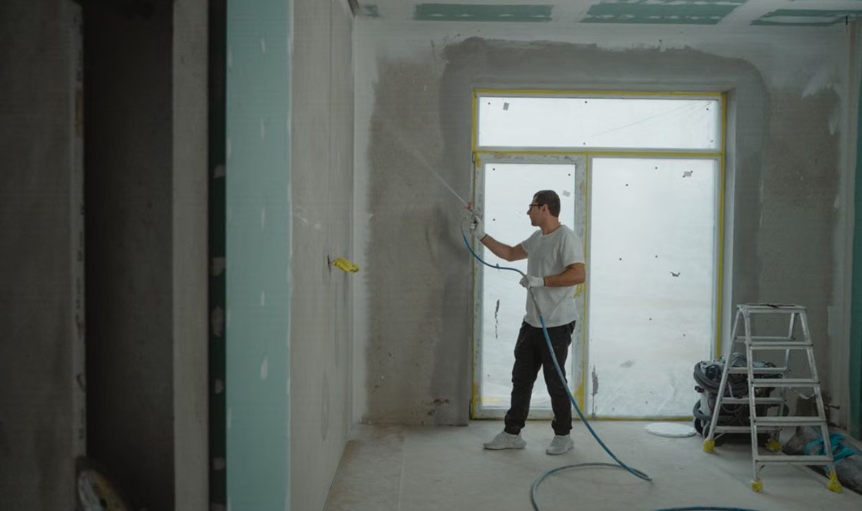 A man sprays paint on a wall while renovating an indoor room with professional equipment.