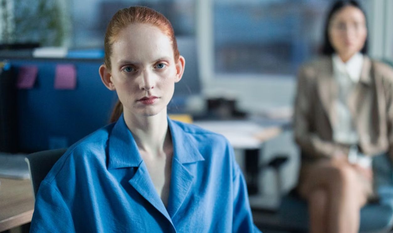 Woman in blue looks focused in modern office with a blurred colleague behind her.