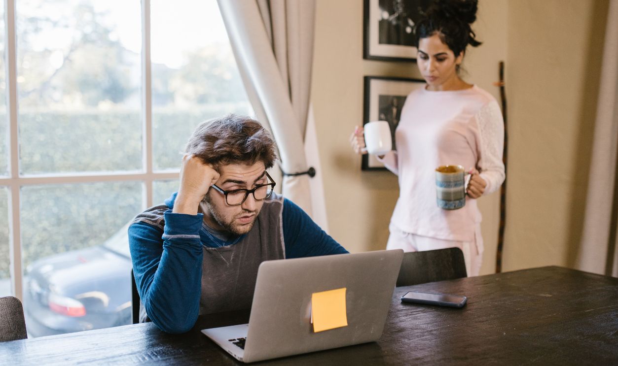 A man appears stressed working on a laptop at home while a woman brings him a drink.