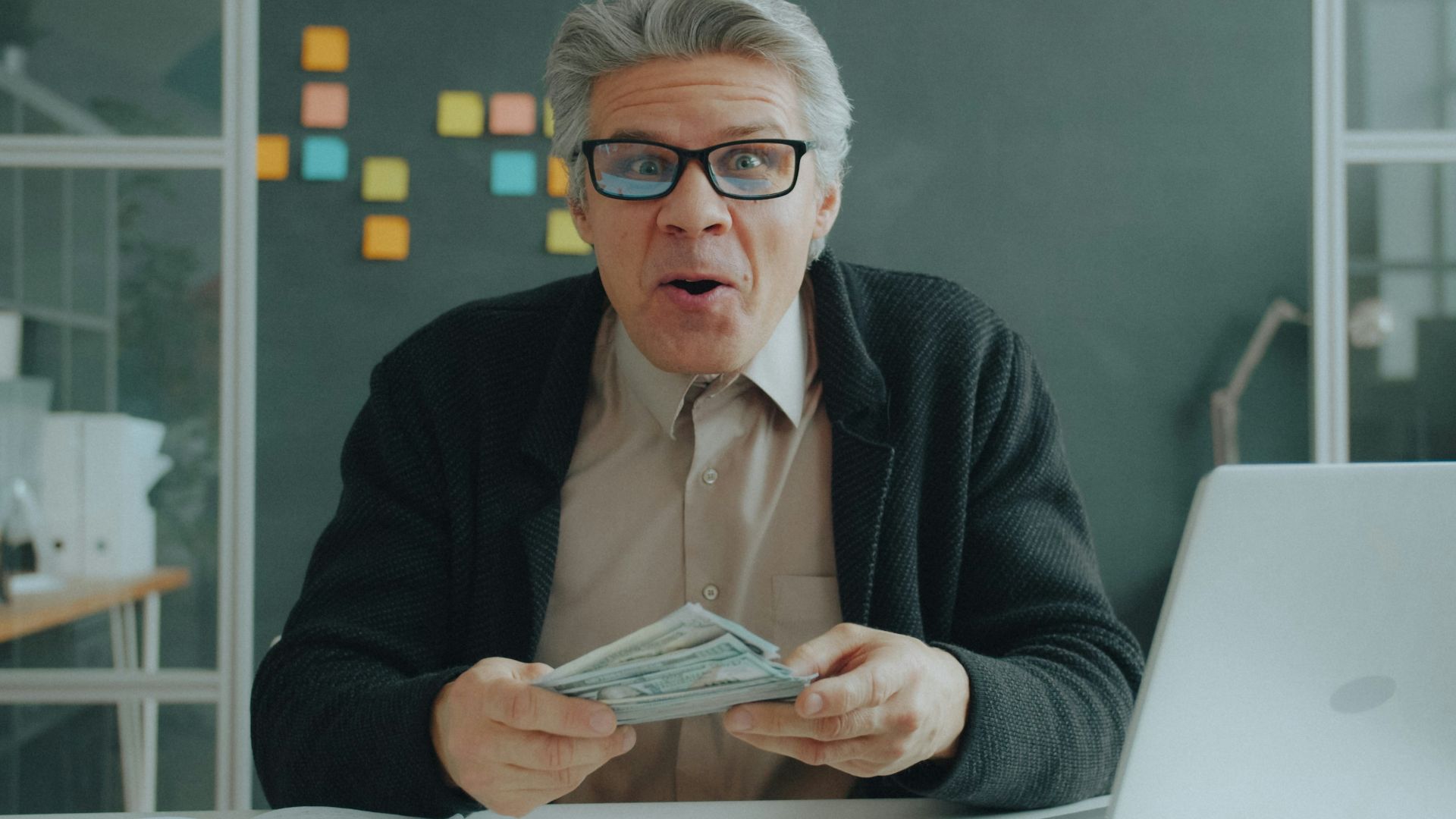 A man with grey hair and glasses holding a stack of cash in an office