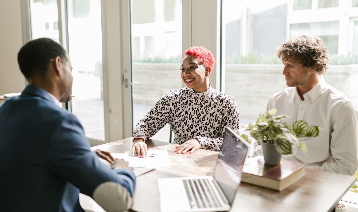 Three professionals meeting in a sunny office discussing finance with smiles.