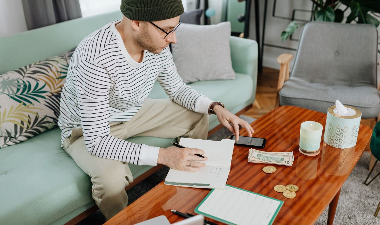 A man in a striped sweater sits on a sofa, writing in a notebook with cash and coins on a wooden table.