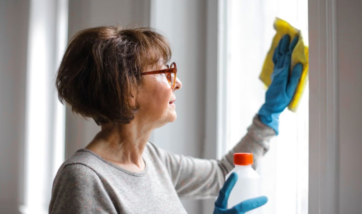 Elderly woman cleansing window indoors using gloves and cleaner.