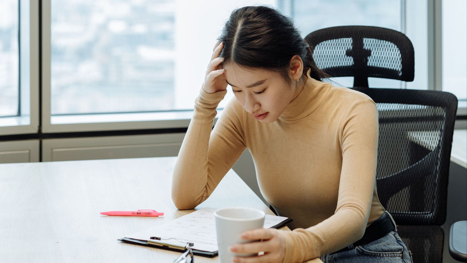Adult woman in office setting, reviewing documents, holding a cup, during daytime.