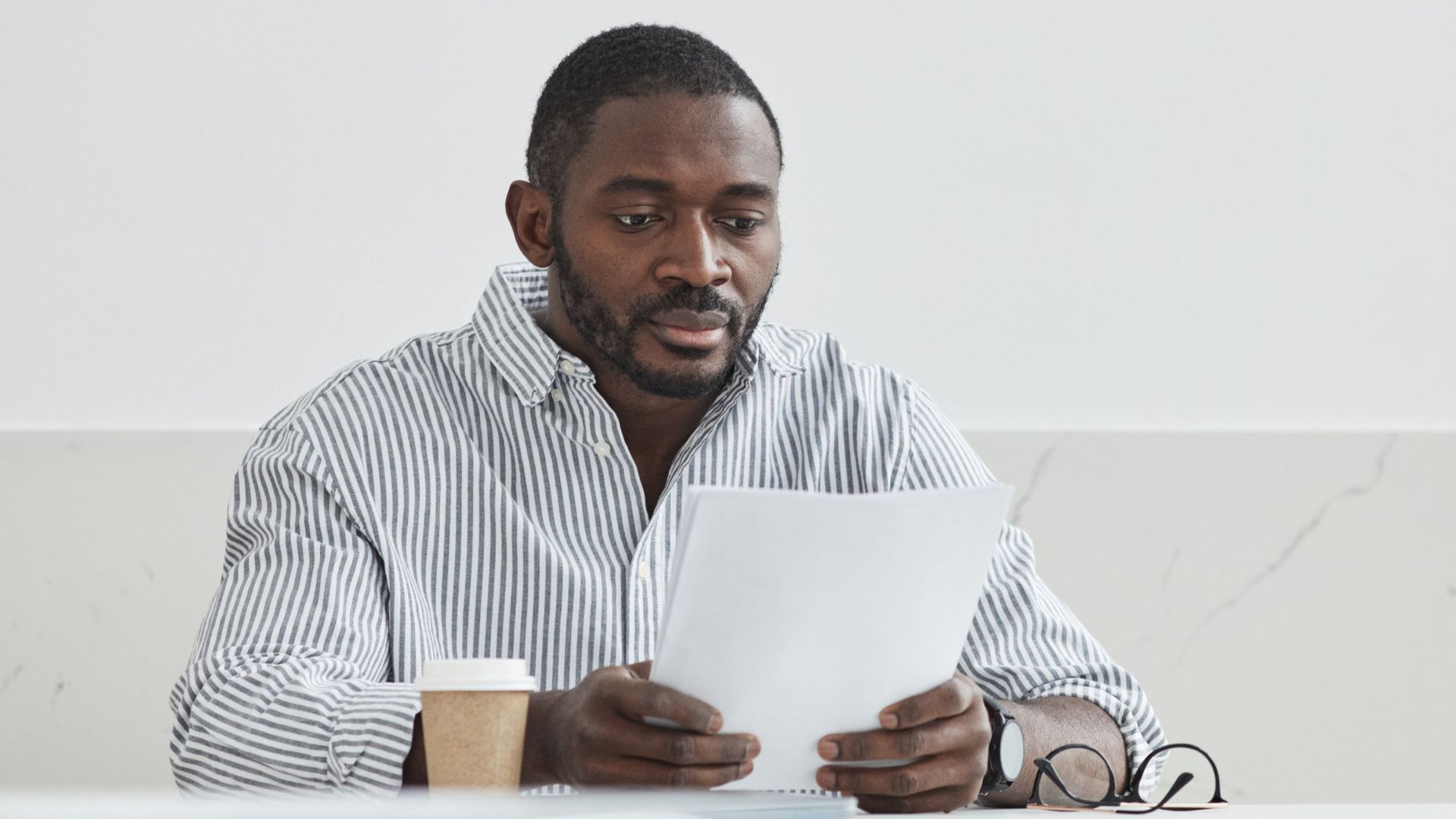 African American man sitting indoors, reading papers with a coffee cup nearby.