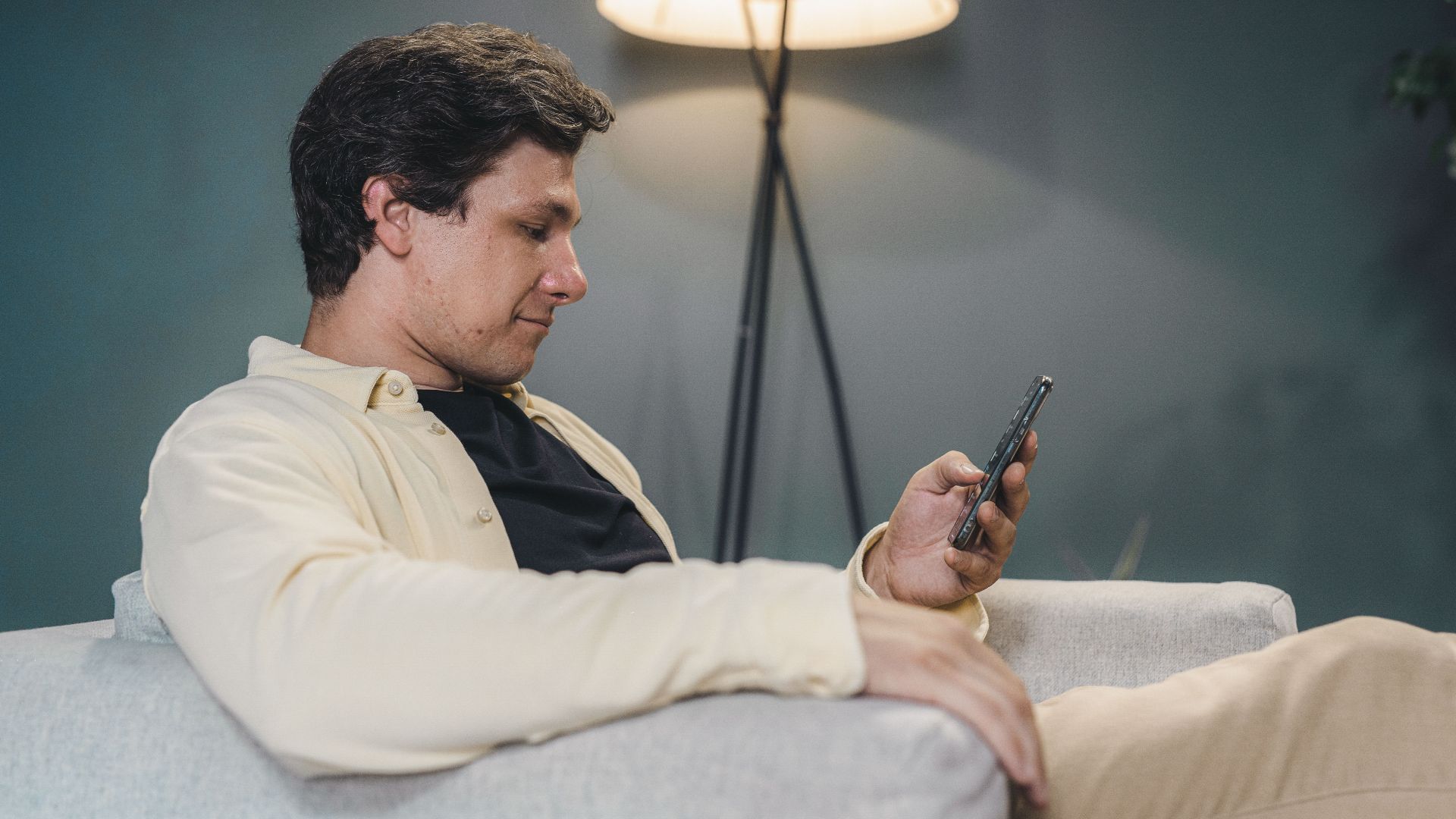 Adult man in modern home interior relaxing on a sofa using his smartphone with warm lighting.