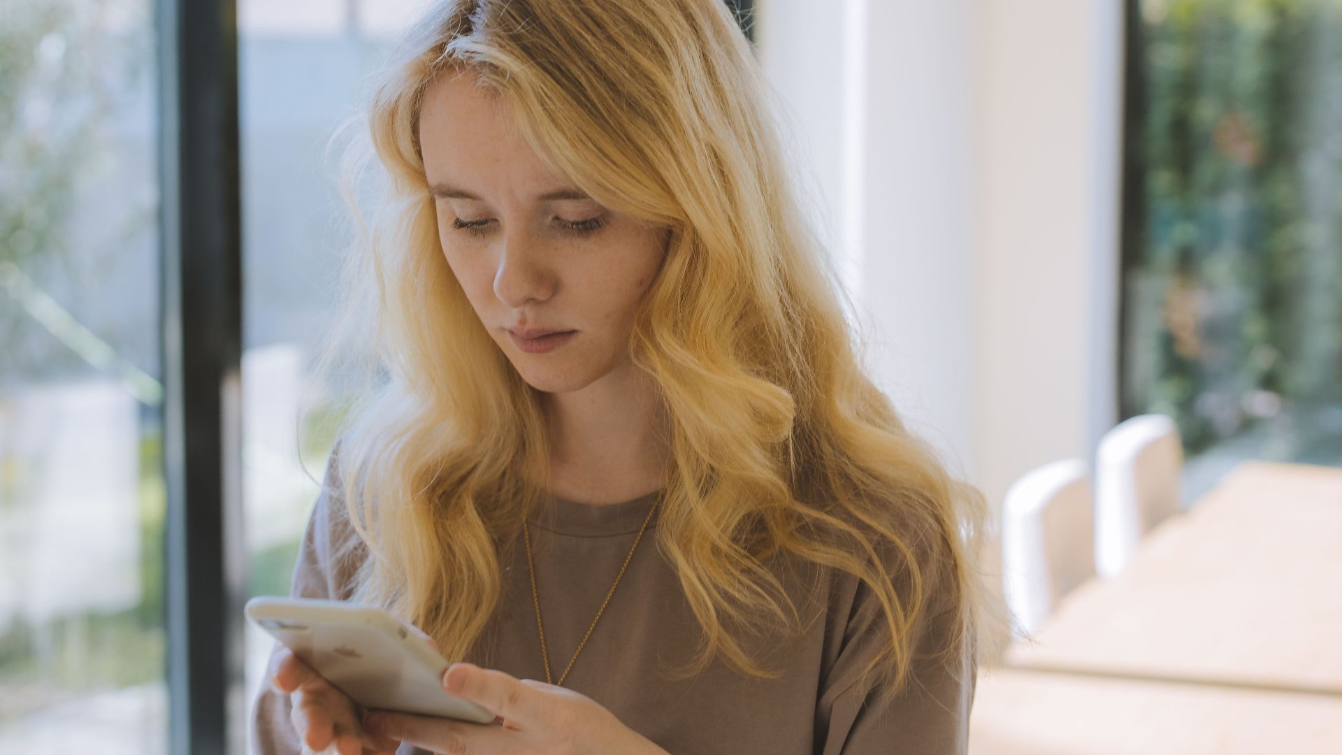 Young woman with blonde hair texting indoors near a large window