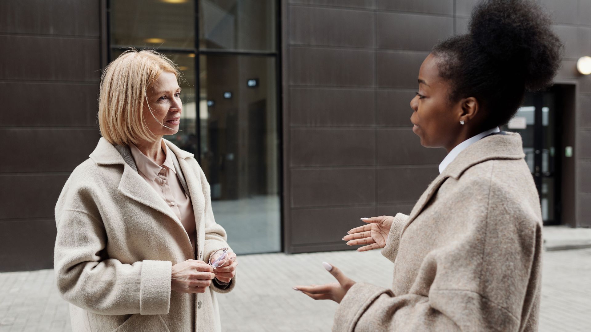 Two businesswomen in coats converse outside a modern office building.