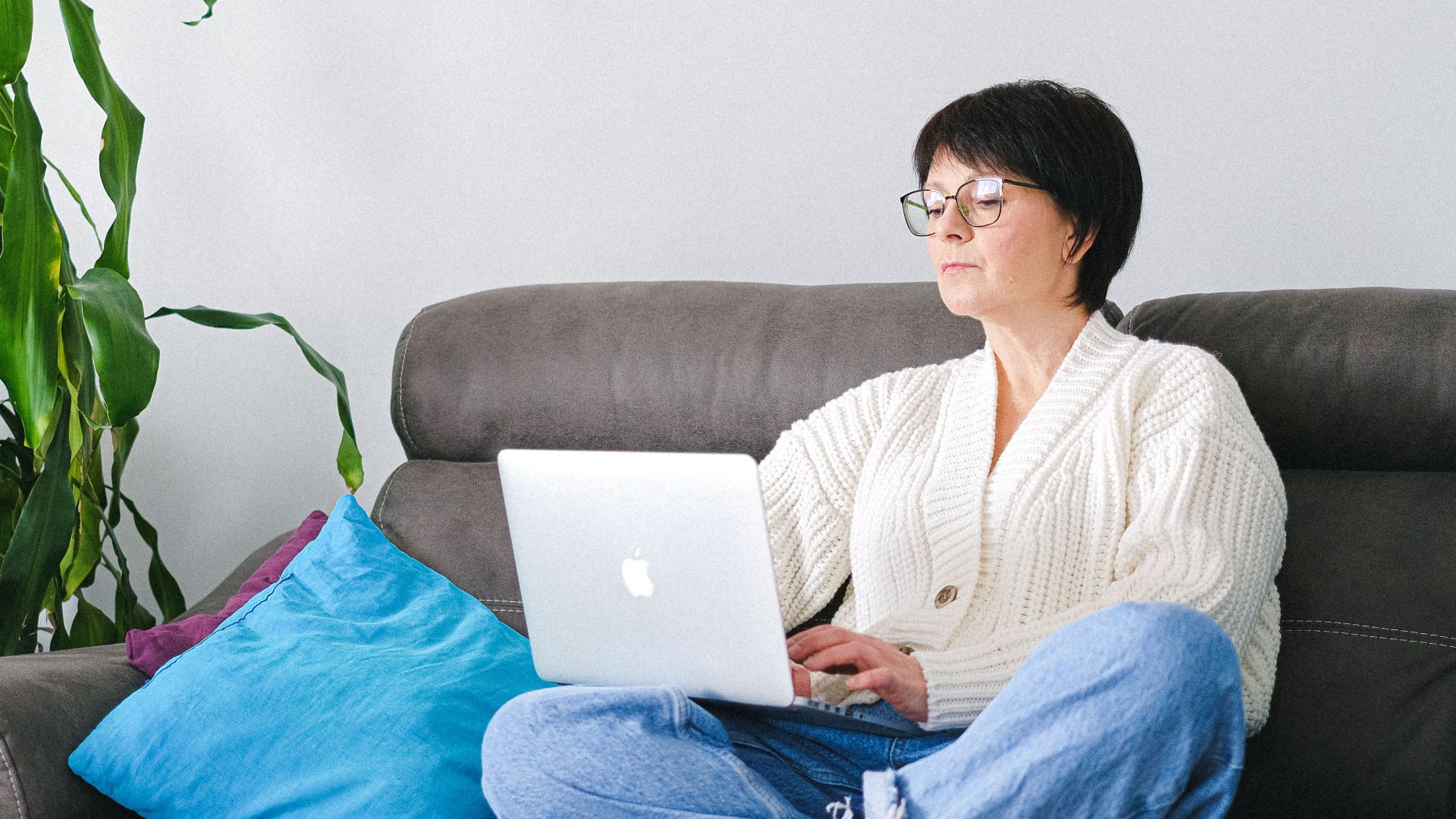 Elderly woman in a cardigan using a laptop while sitting on a couch surrounded by home decor.