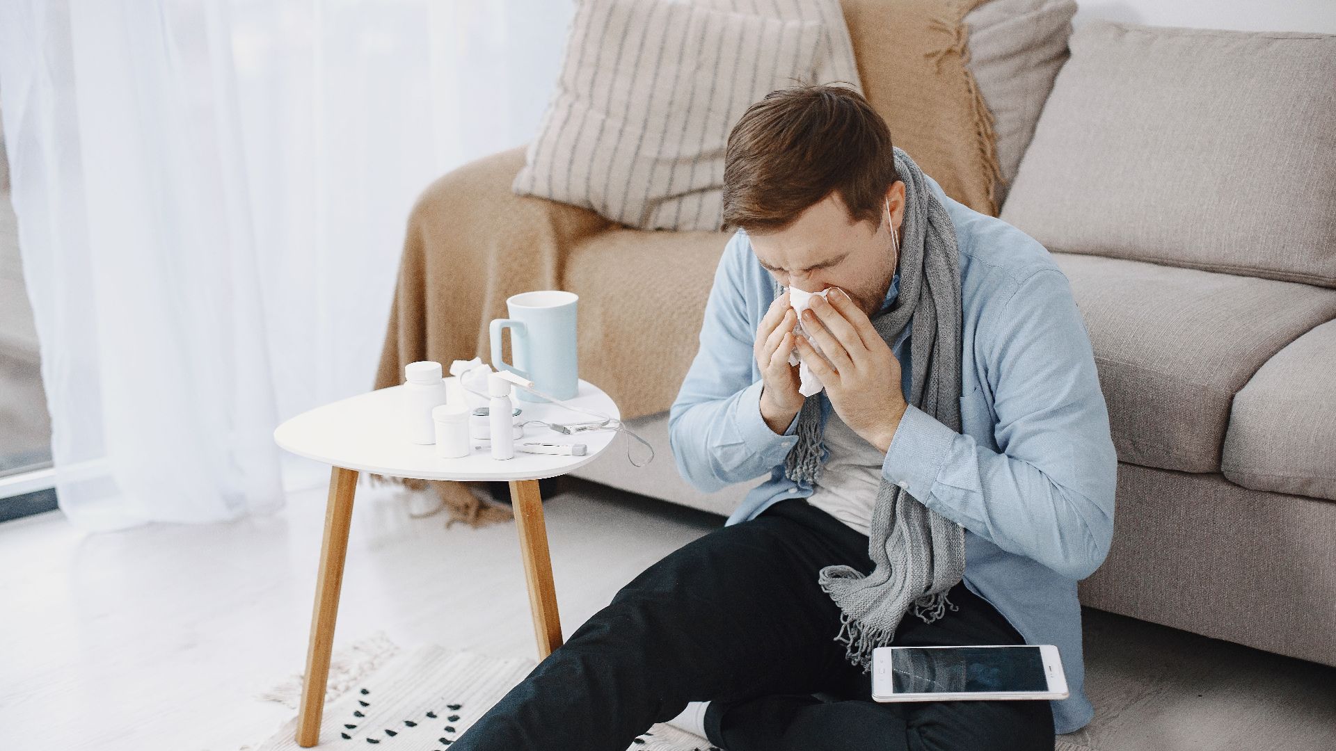 A man in a living room sneezing into a tissue, surrounded by a table, sofa, and digital tablet.