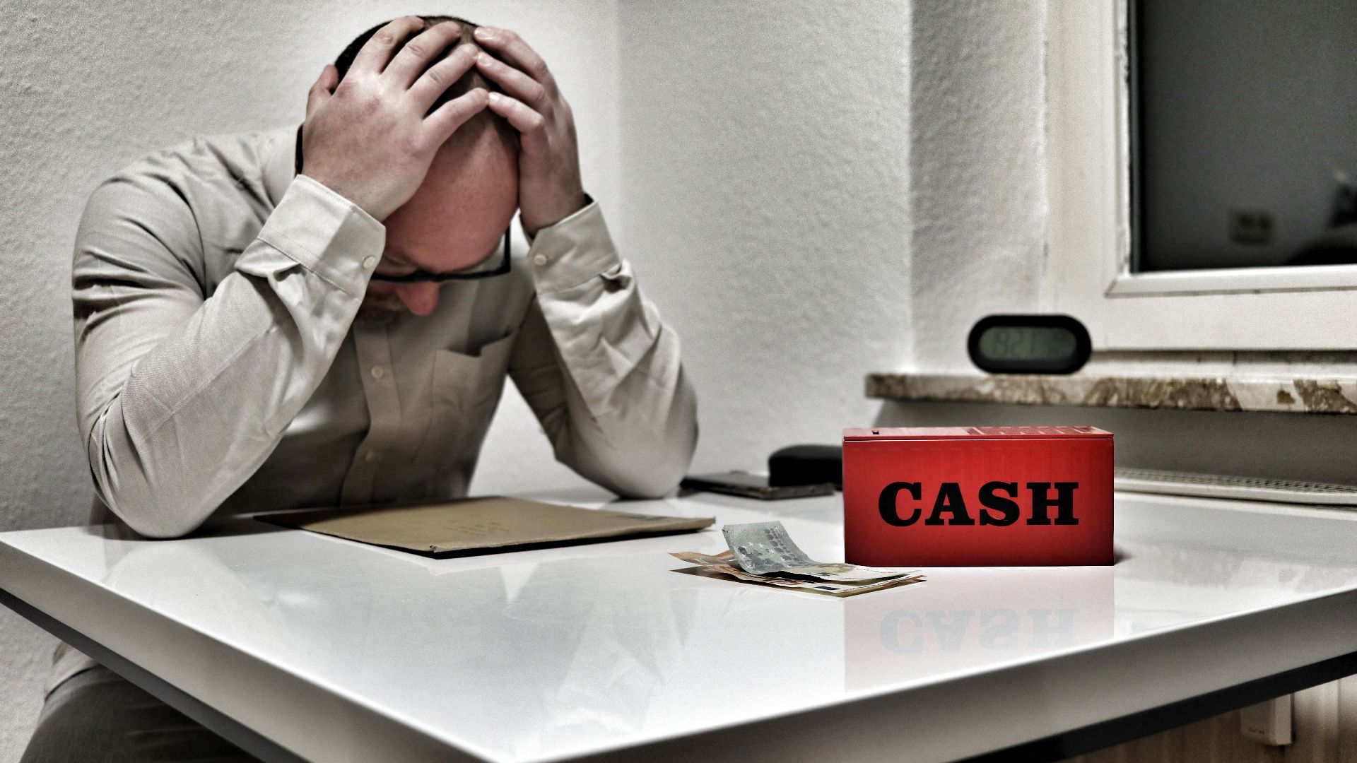 Bald man holding head in despair at desk with cashbox and money. Indoor office stress concept.