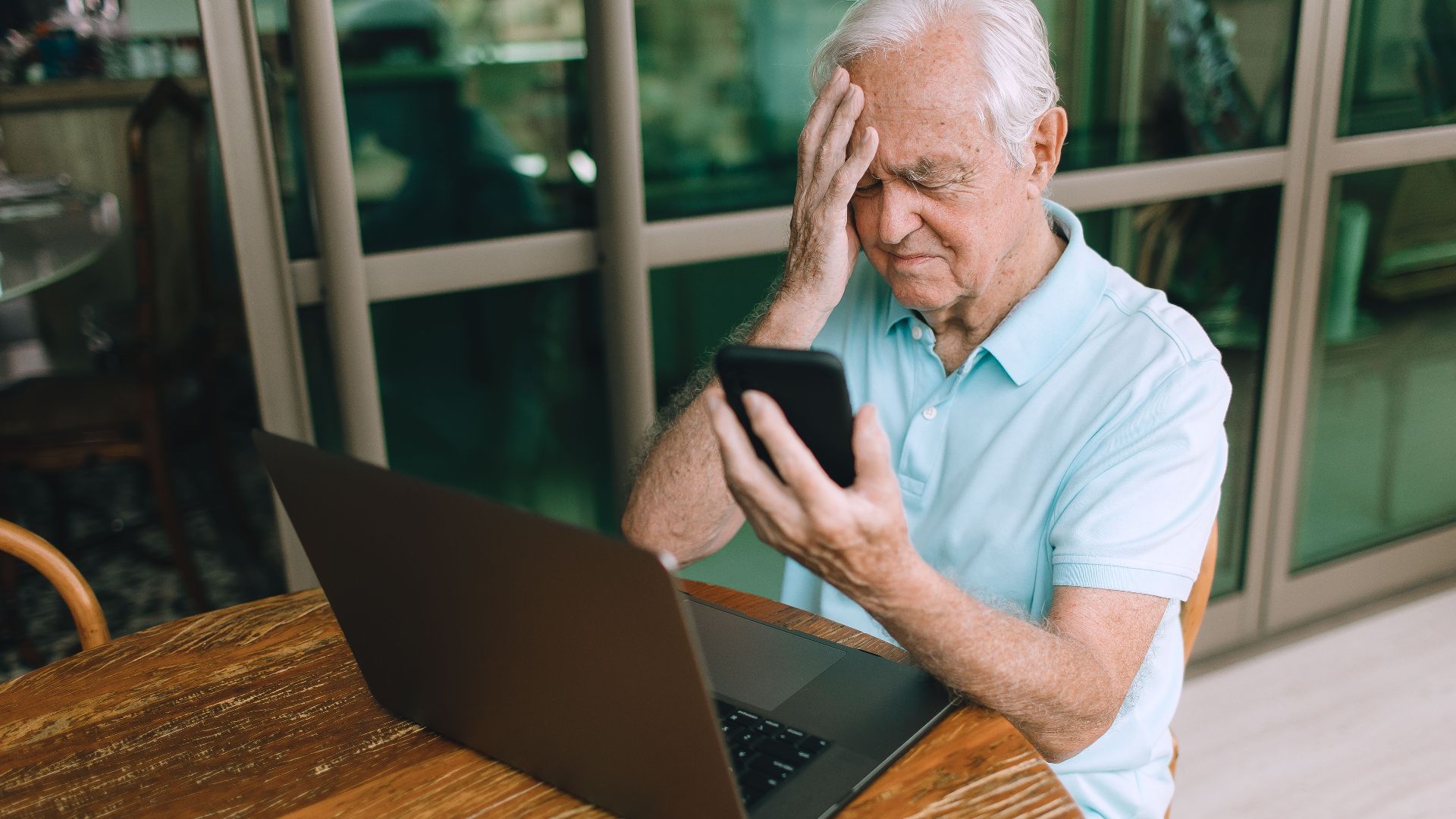 An elderly man in a blue shirt looks troubled while holding a smartphone and sitting at a table with a laptop.