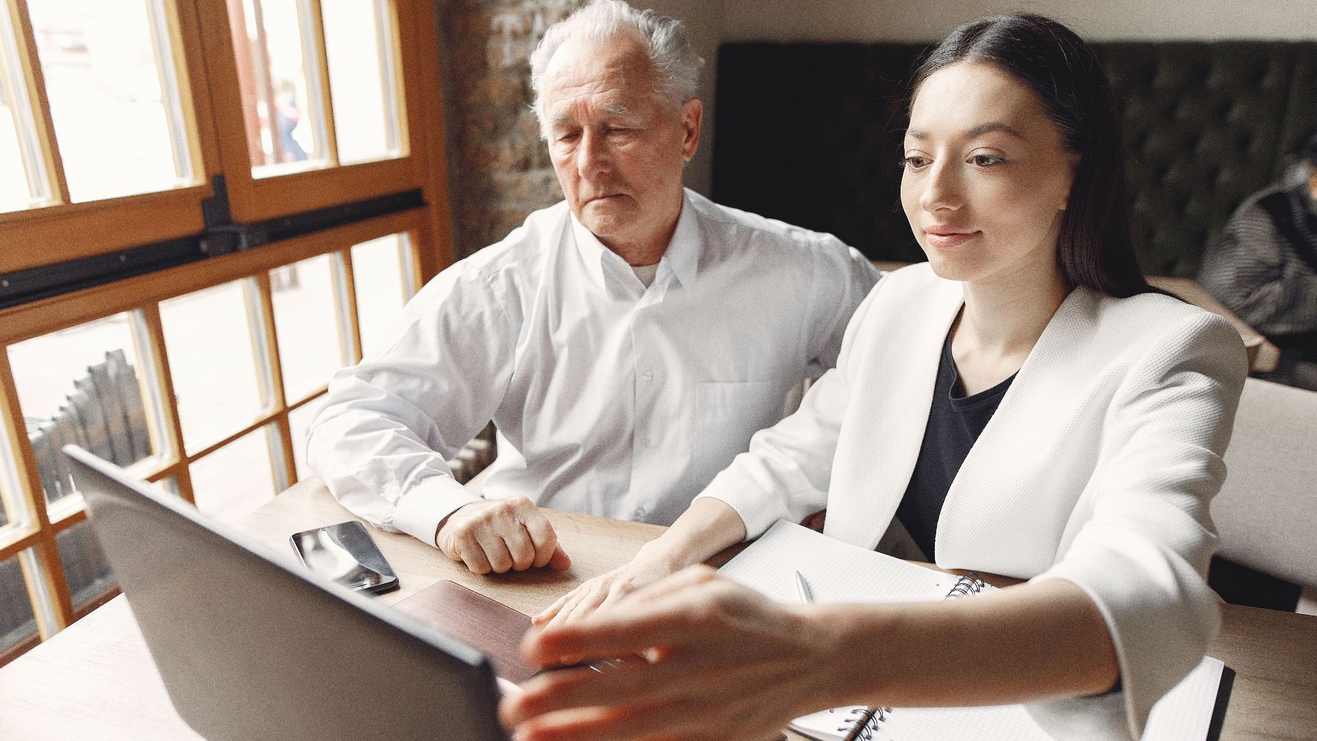 Modern adult woman with senior male executive browsing laptop and checking notes while working remotely in creative workspace near window and looking at screen