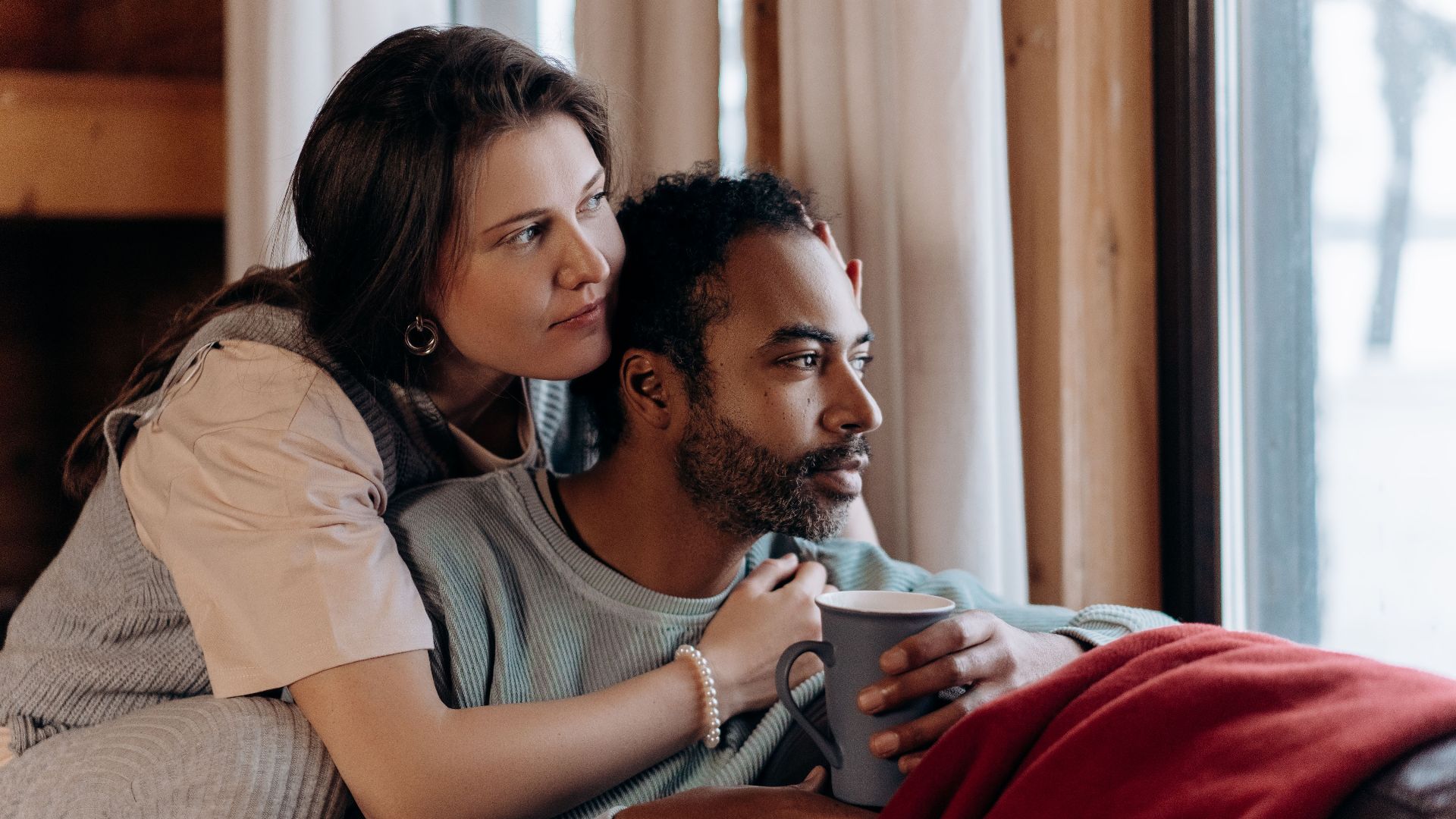 A multiethnic couple enjoys a cozy moment indoors, sitting by a window with a warm embrace.