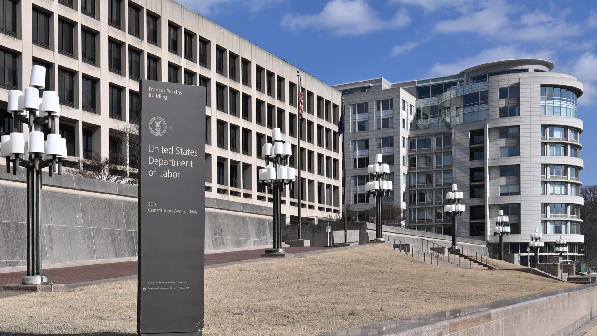 A sign on Constitution Ave for the United States Department of Labor with the Frances Perkins building behind it. 200 Constitution Avenue NW, Washington, DC 20004.