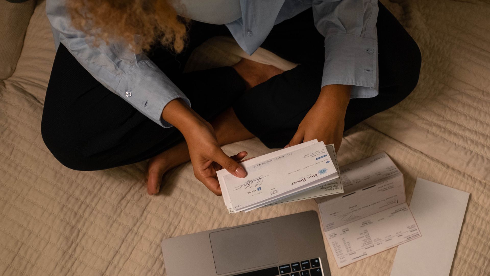 Overhead view of woman organizing finances on bed with laptop and checks.