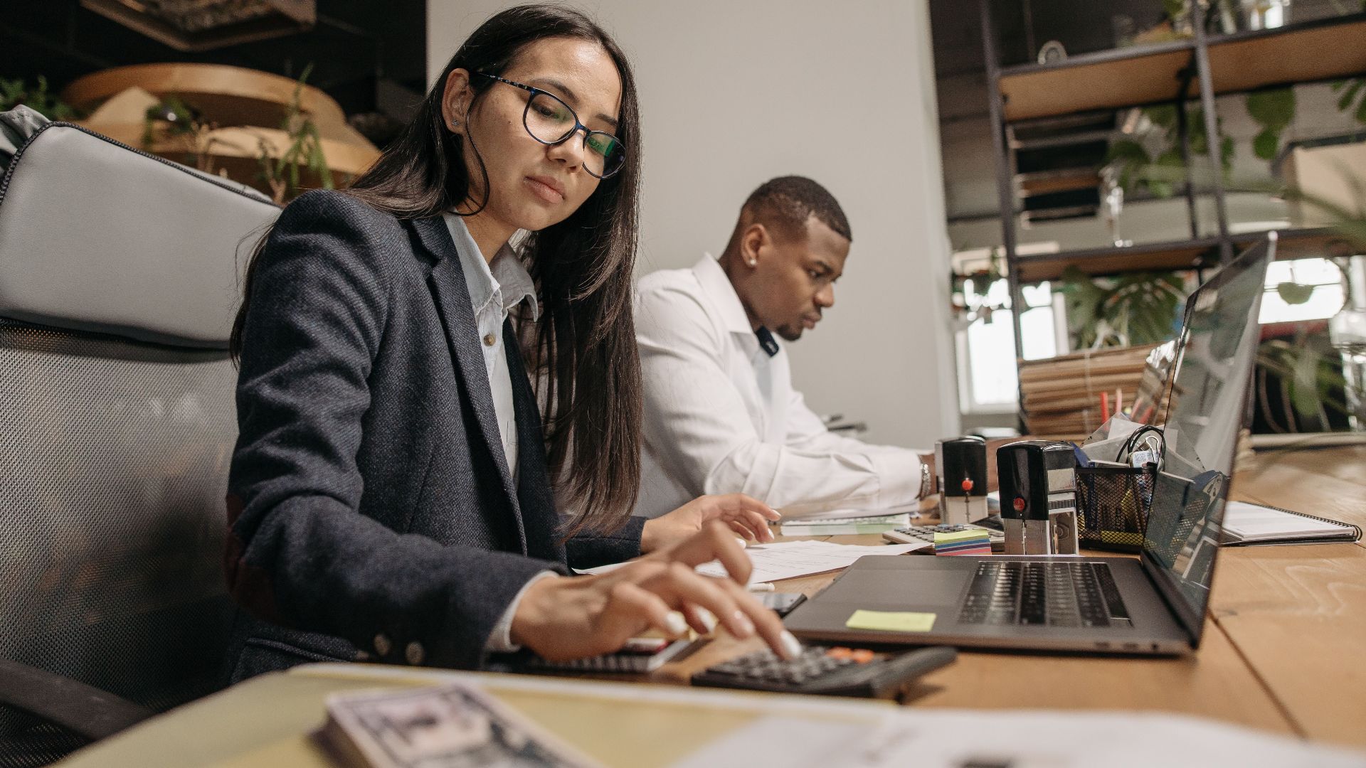 A busy office scene with two professionals working on laptops and calculators.