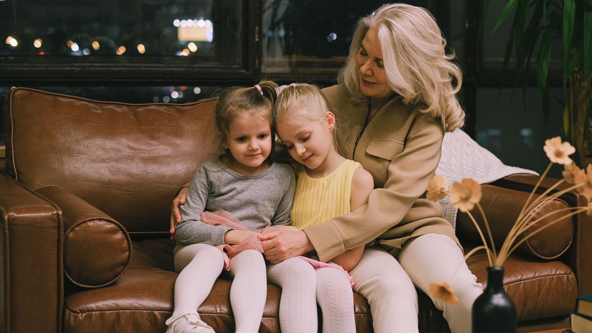 Grandmother lovingly embraces two granddaughters on a cozy leather sofa indoors.