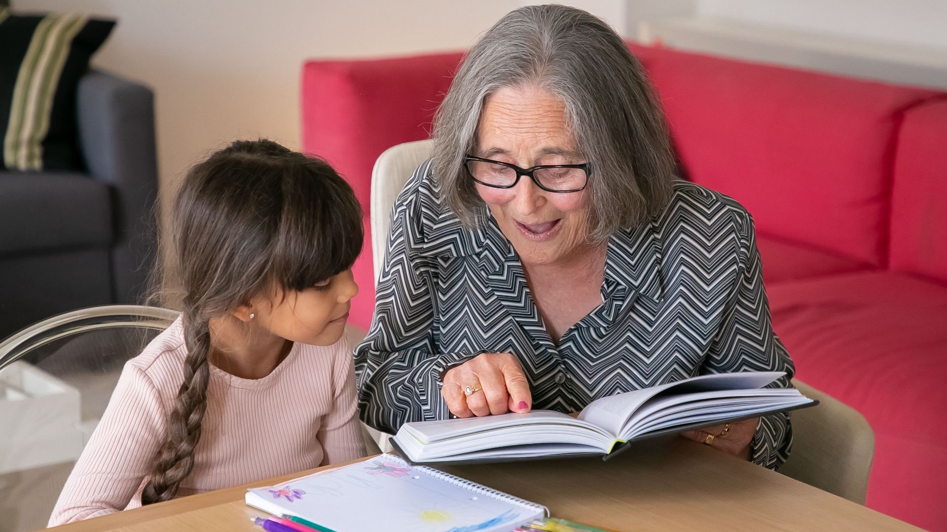 A joyful grandmother reading a book with her granddaughter indoors, bonding over learning.