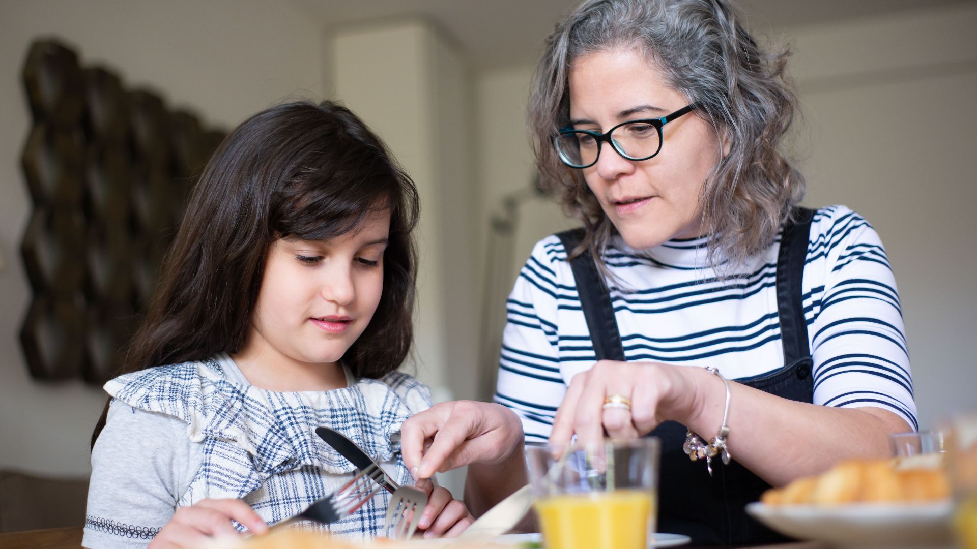 Grandmother guides young girl in table manners during breakfast at home.