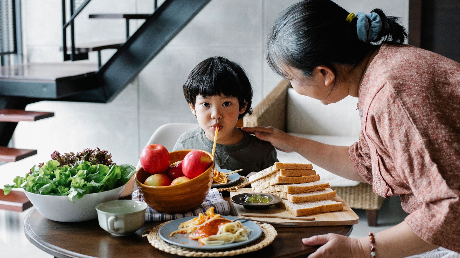Cute funny Asian boy sucking spaghetti while gathering at dining table with positive grandmother in light kitchen