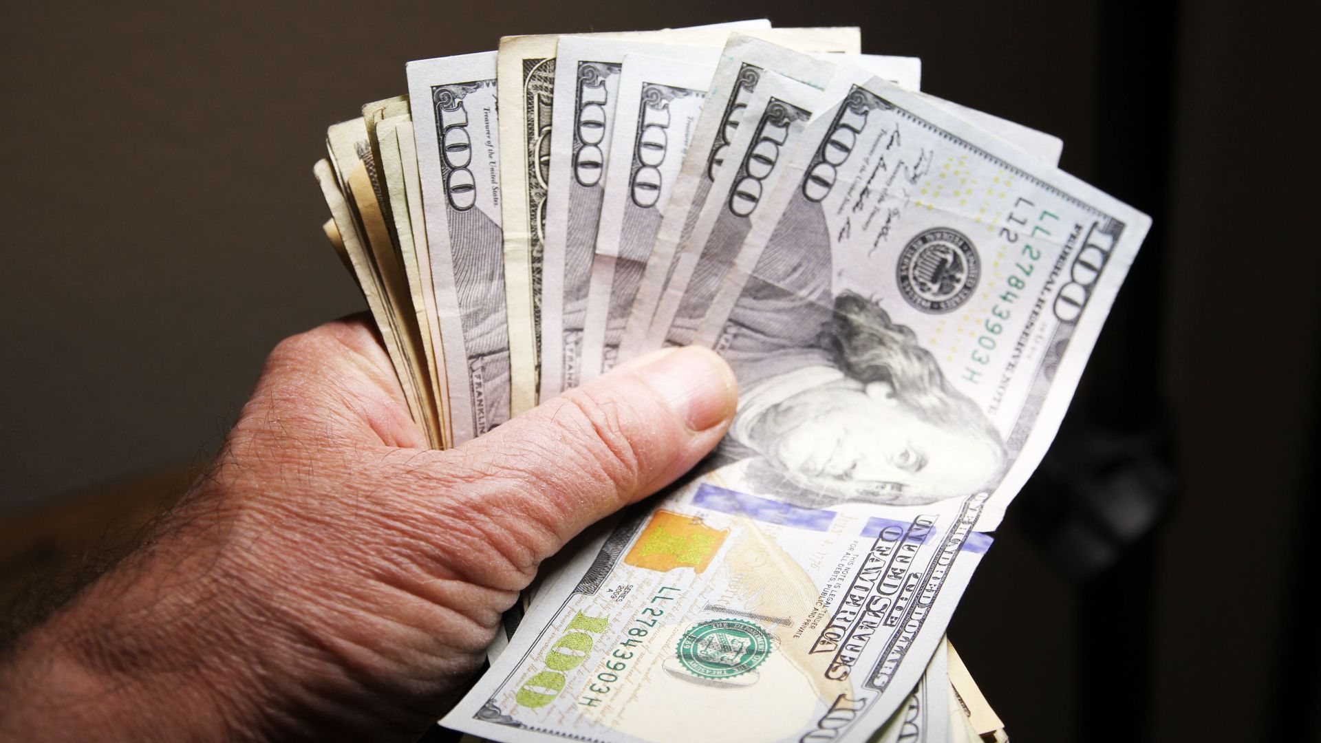 Close-up of a hand holding multiple hundred dollar bills on a dark background.