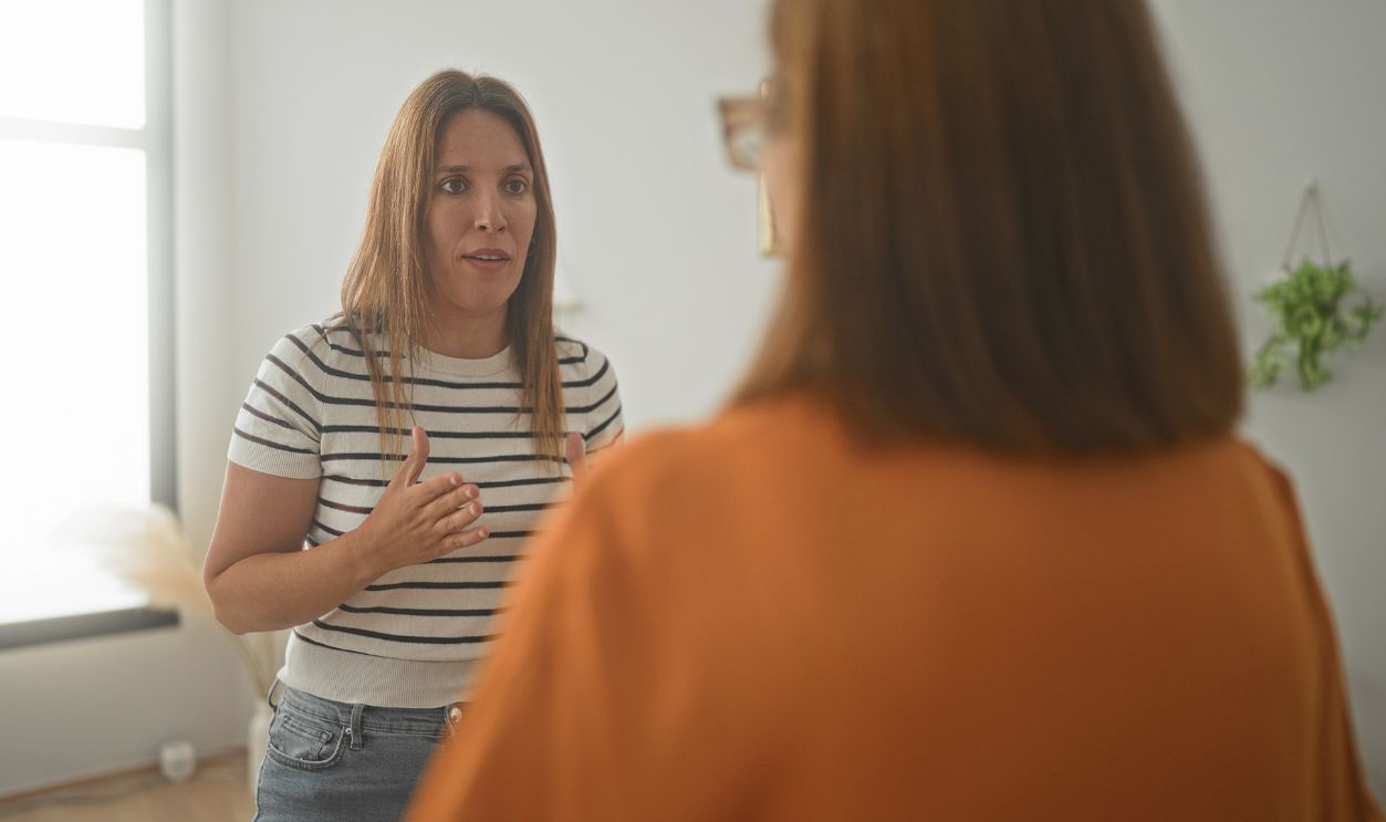Woman gesturing with hand while speaking to another woman in a livingroom, palms visible and expressive body language during a domestic conversation; family tension.