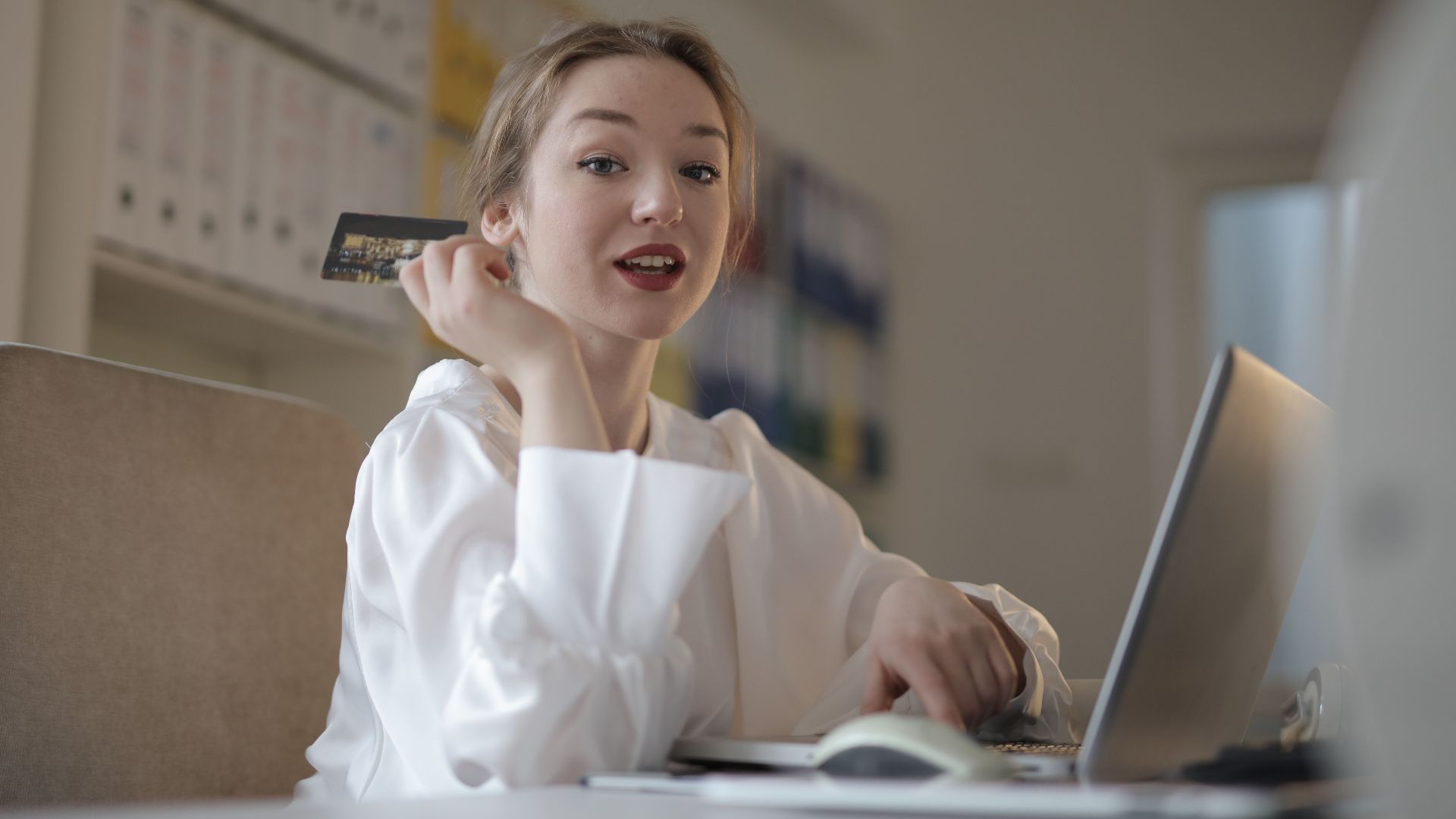 Low angle side view of positive young female entrepreneur in formal wear using laptop and performing online payment with credit card while sitting at table at workplace