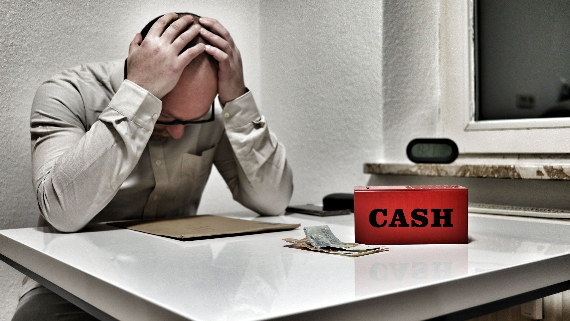 Bald man holding head in despair at desk with cashbox and money. Indoor office stress concept.