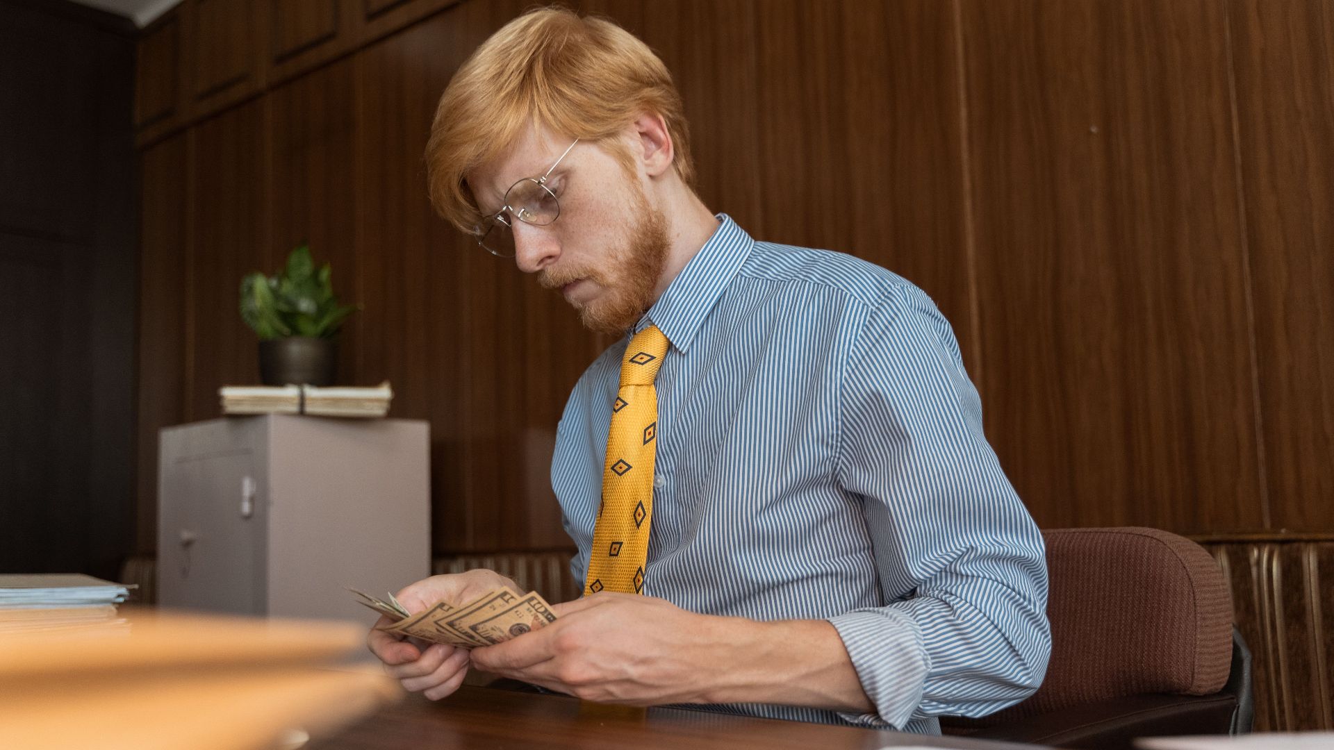 Serious businessman counting cash at his desk in an office environment with wood paneling.