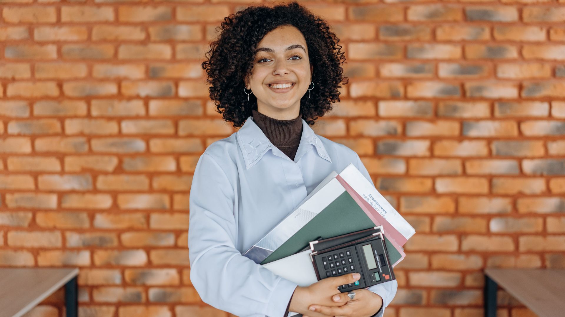 Cheerful woman with curly hair holding folders and a calculator against a brick wall.