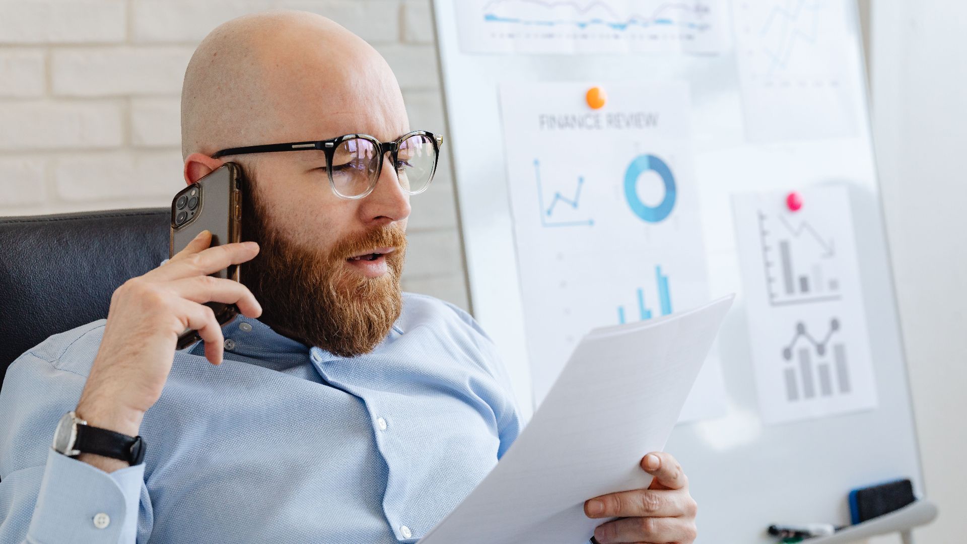 Bald man with beard holding smartphone and papers, reviewing financial charts in office.