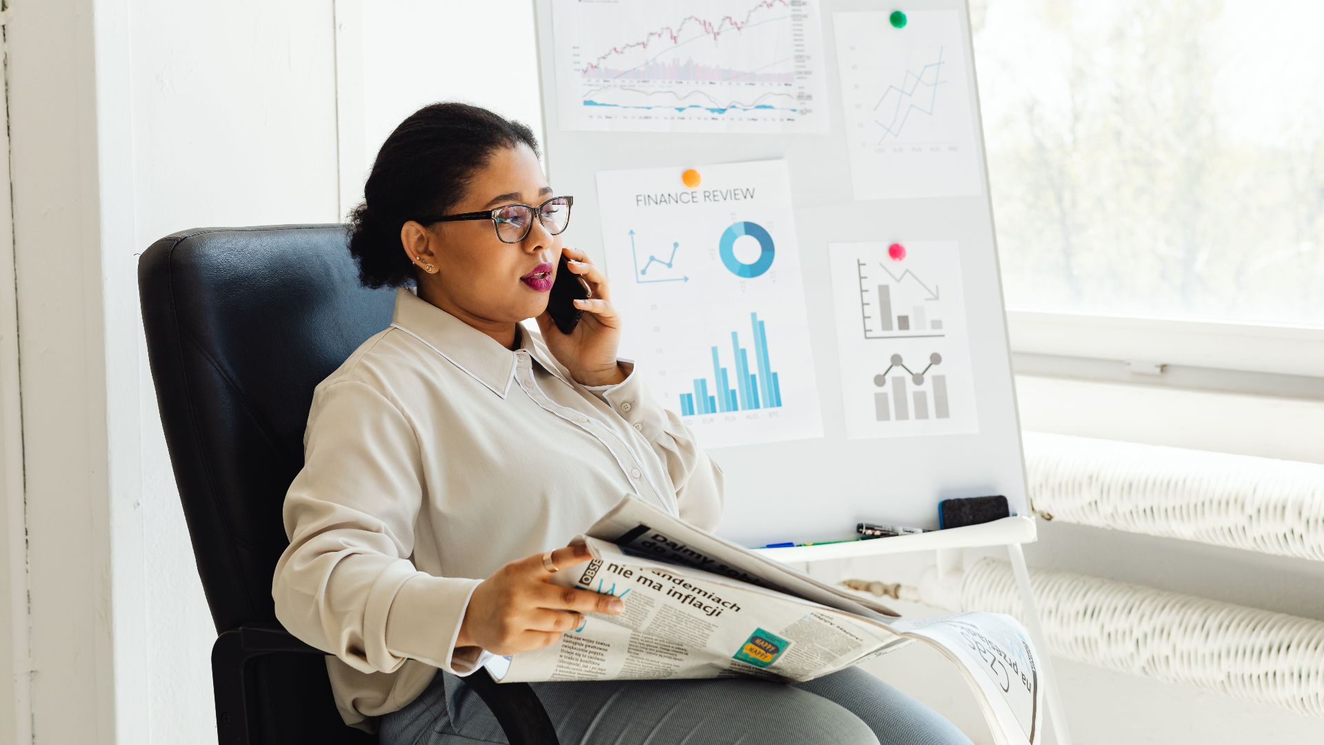 Businesswoman multitasking with phone and newspaper in office, examining financial charts.