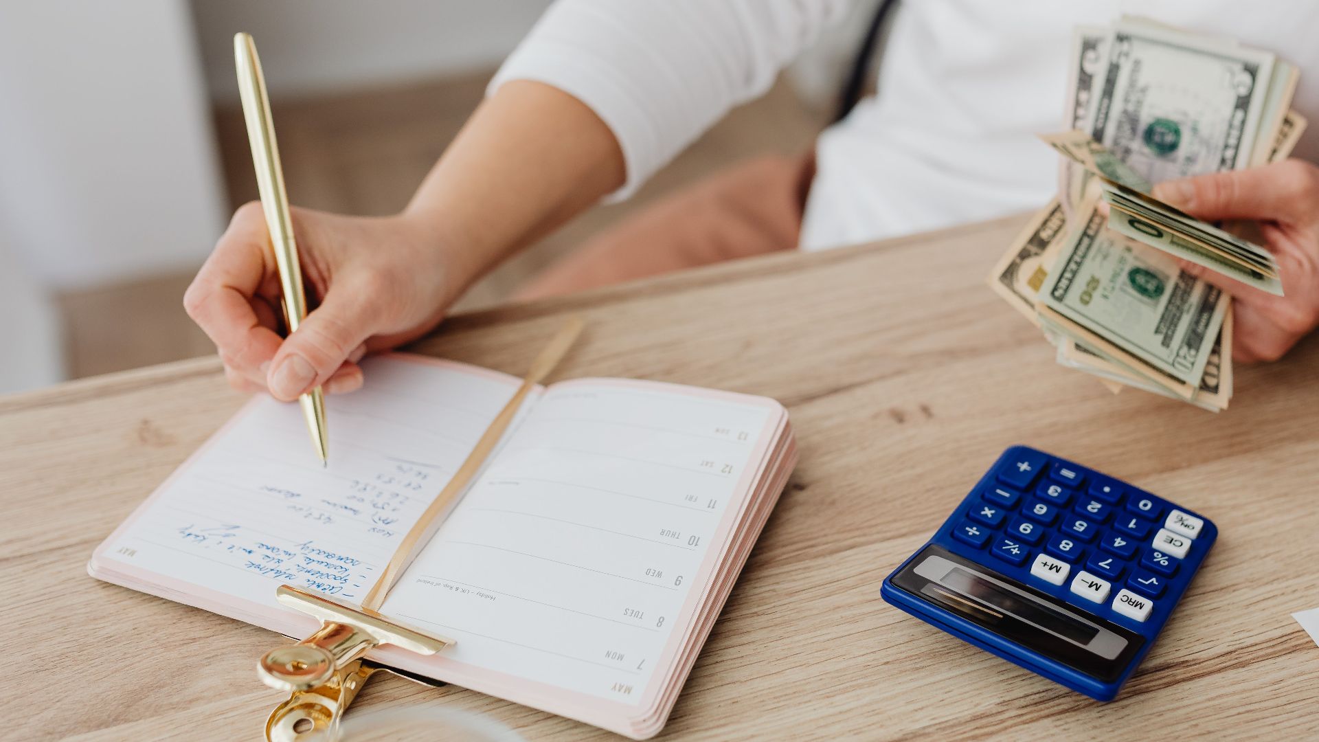 Adult holding cash and writing in planner while using a calculator at home.
