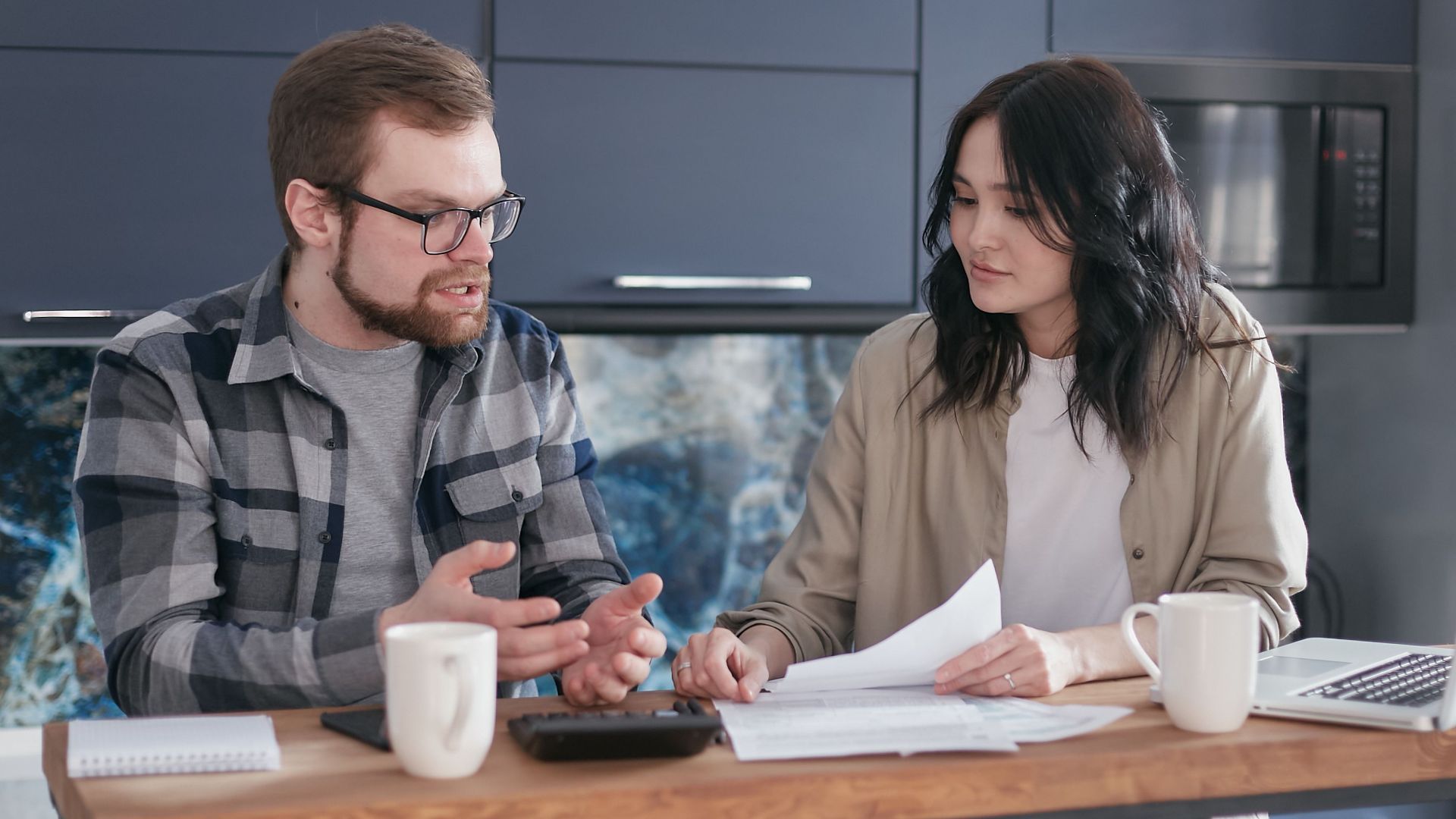 A couple engaged in a financial discussion, sitting at a kitchen table with papers and a laptop.