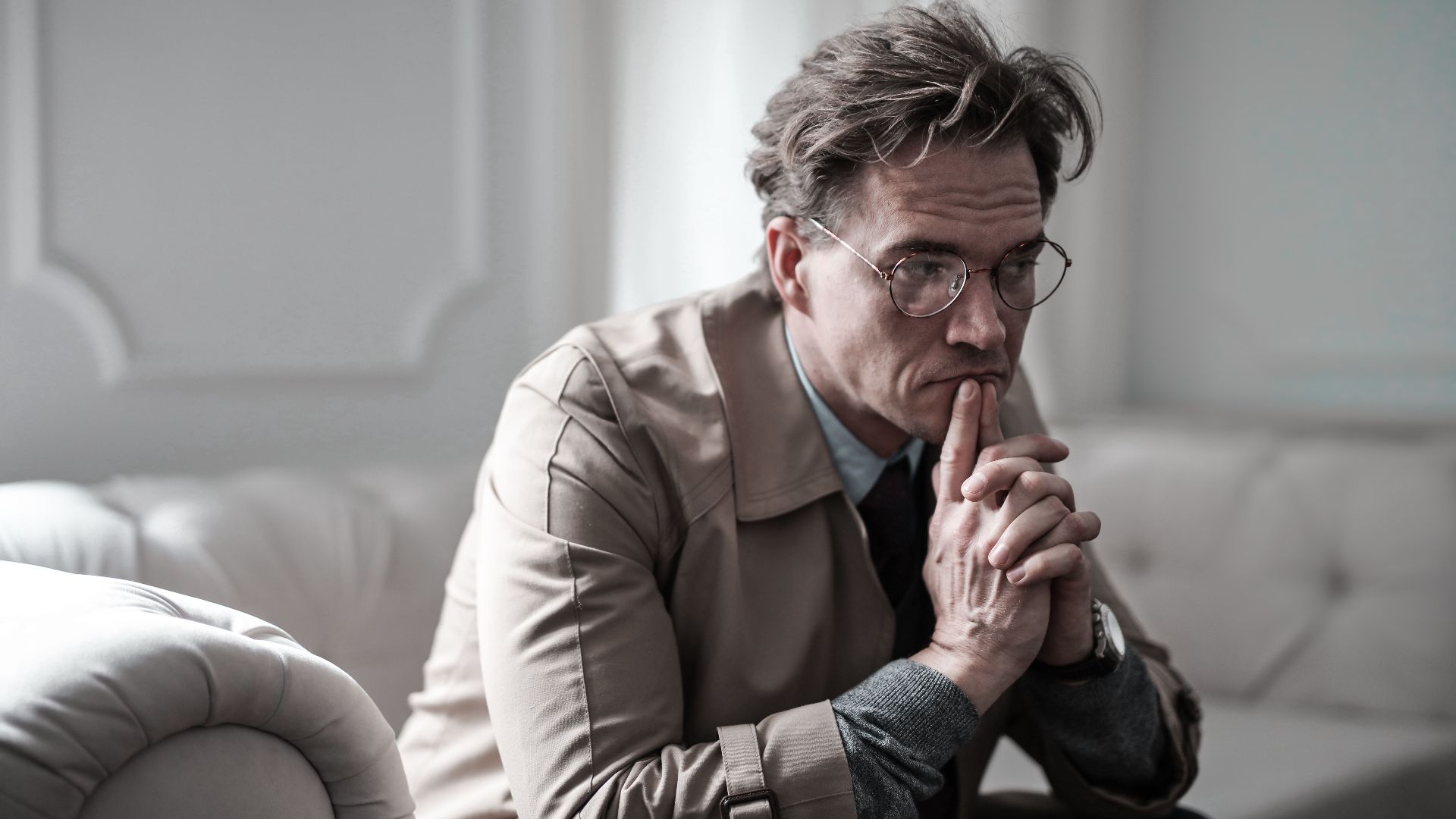 A thoughtful man wearing glasses sits on a sofa in a classic interior, lost in deep contemplation.