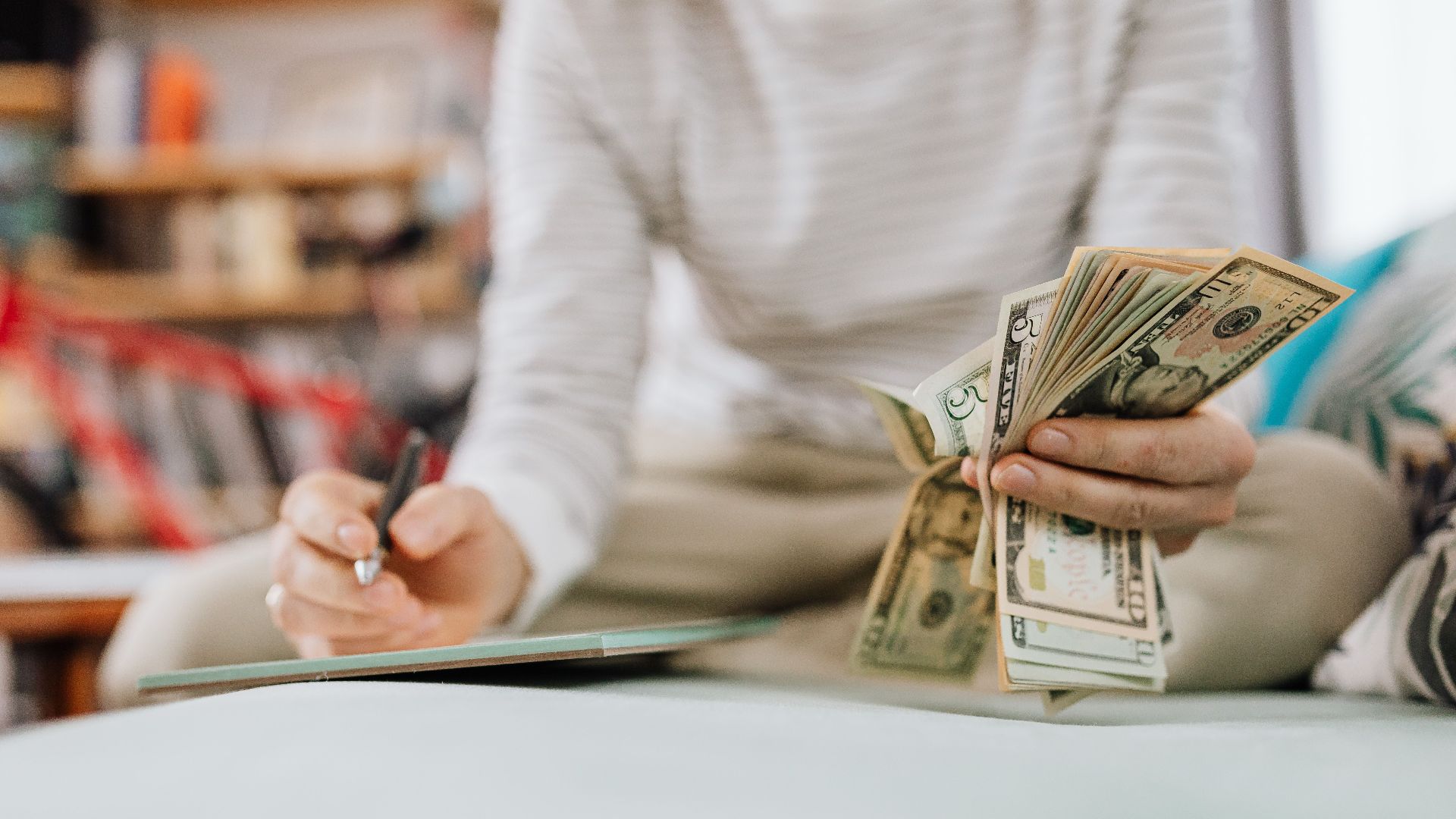 Individual counting money with pen in hand inside a cozy room setting.