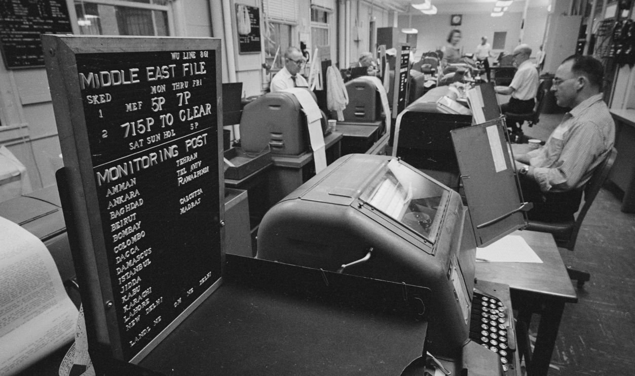 View of teletype machines and workers in a US Information Agency (USIA) office, Washington DC, January 25, 1967. A sign at for reads, in part, 'Middle East File.'