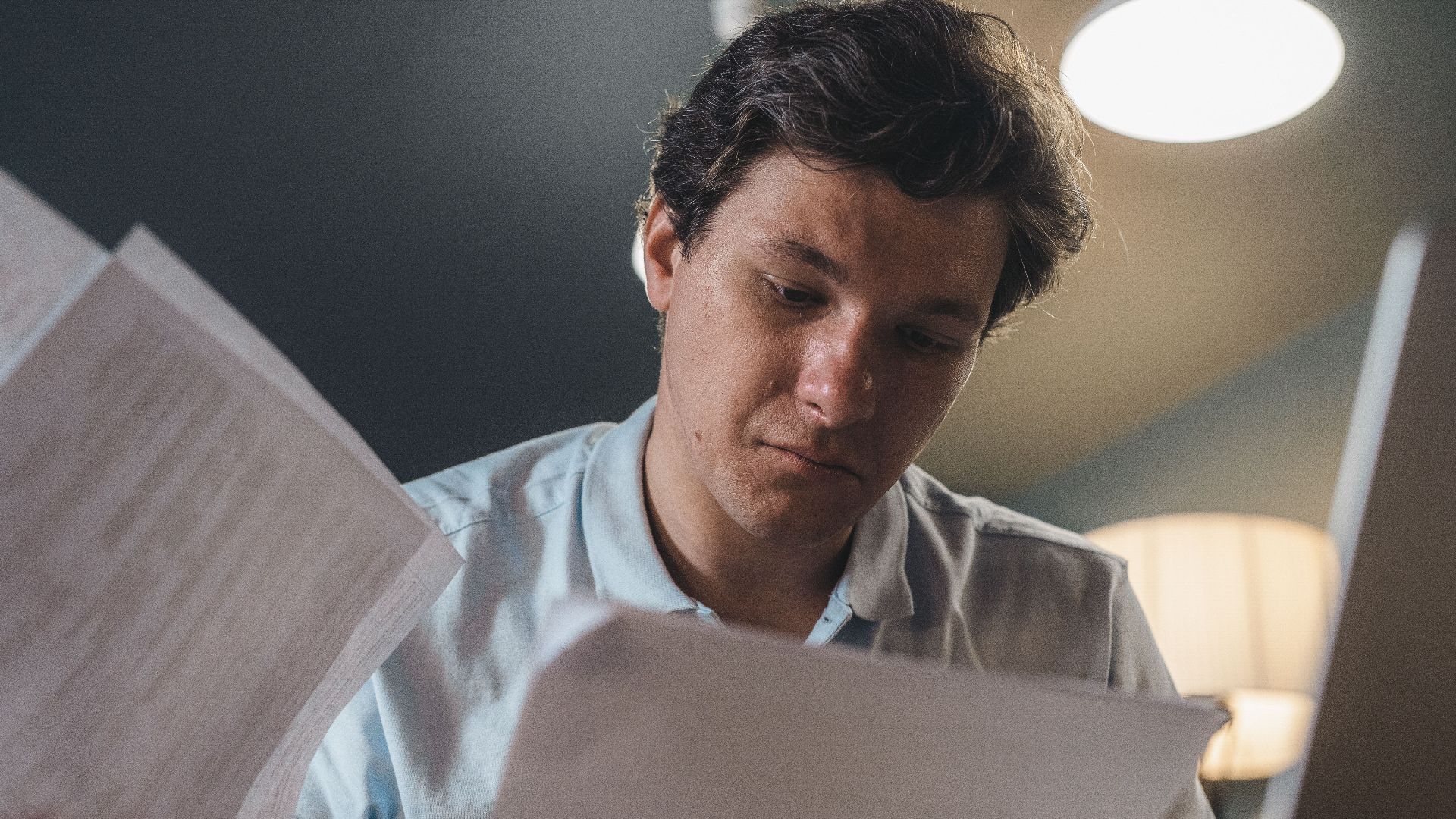 Serious man wearing a light blue shirt intently reading printed papers indoors under warm lighting.