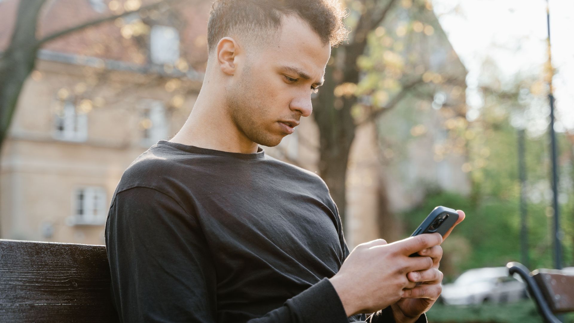 A young man sitting on a park bench using his smartphone during a sunny day.