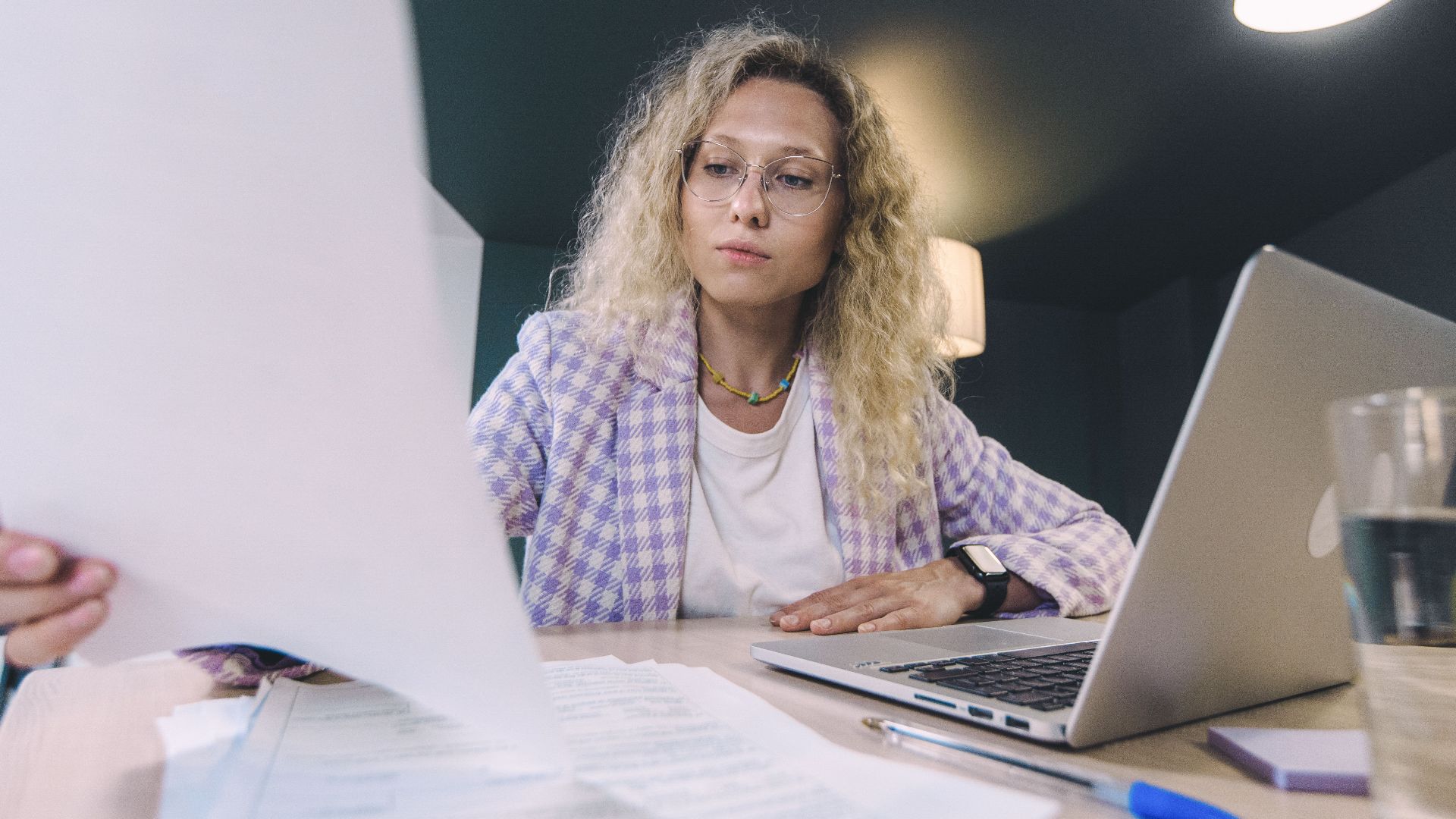 Woman in an office environment reviewing documents with focus, surrounded by technology.
