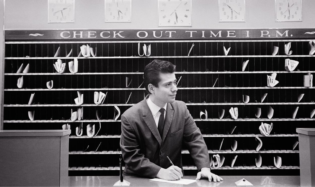 1960s Hotel clerk at front desk message slots check out time 1 pm clocks various world times empire hotel new york city USA.