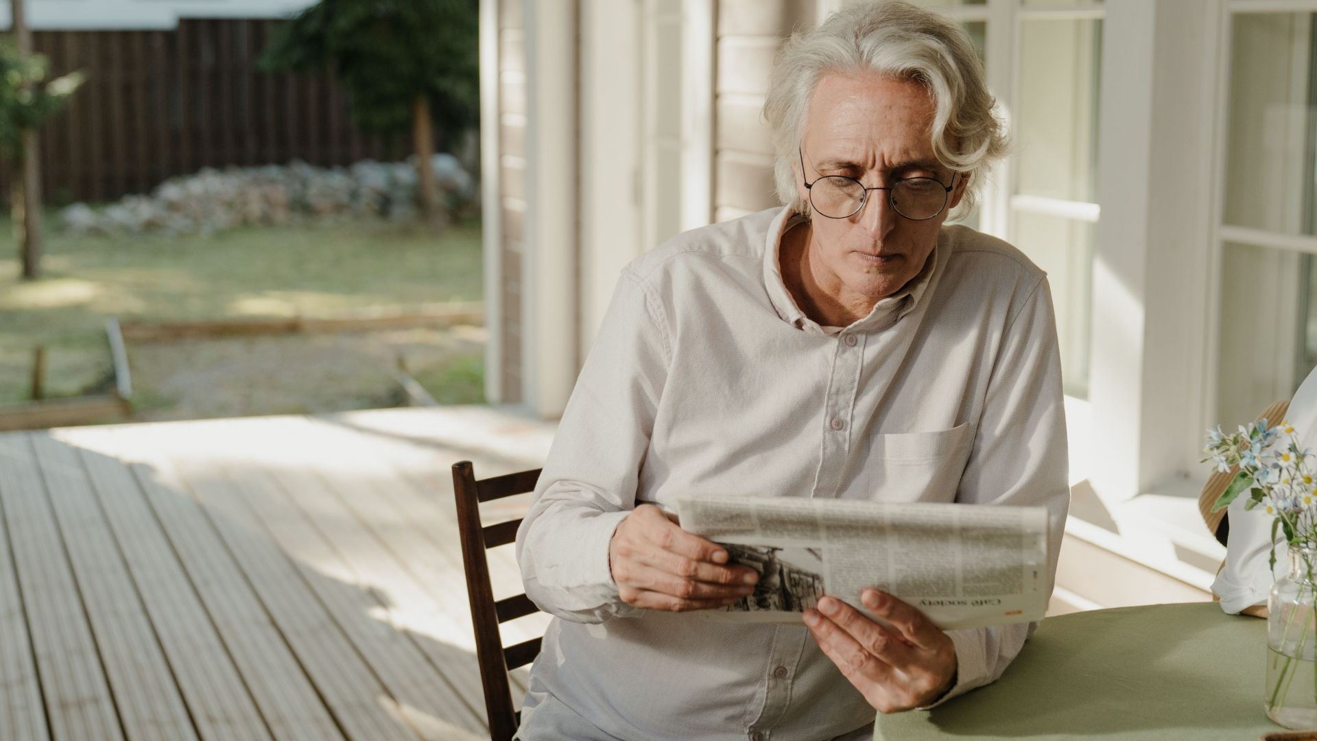 Elderly man with gray hair reading the newspaper on a sunny outdoor veranda.
