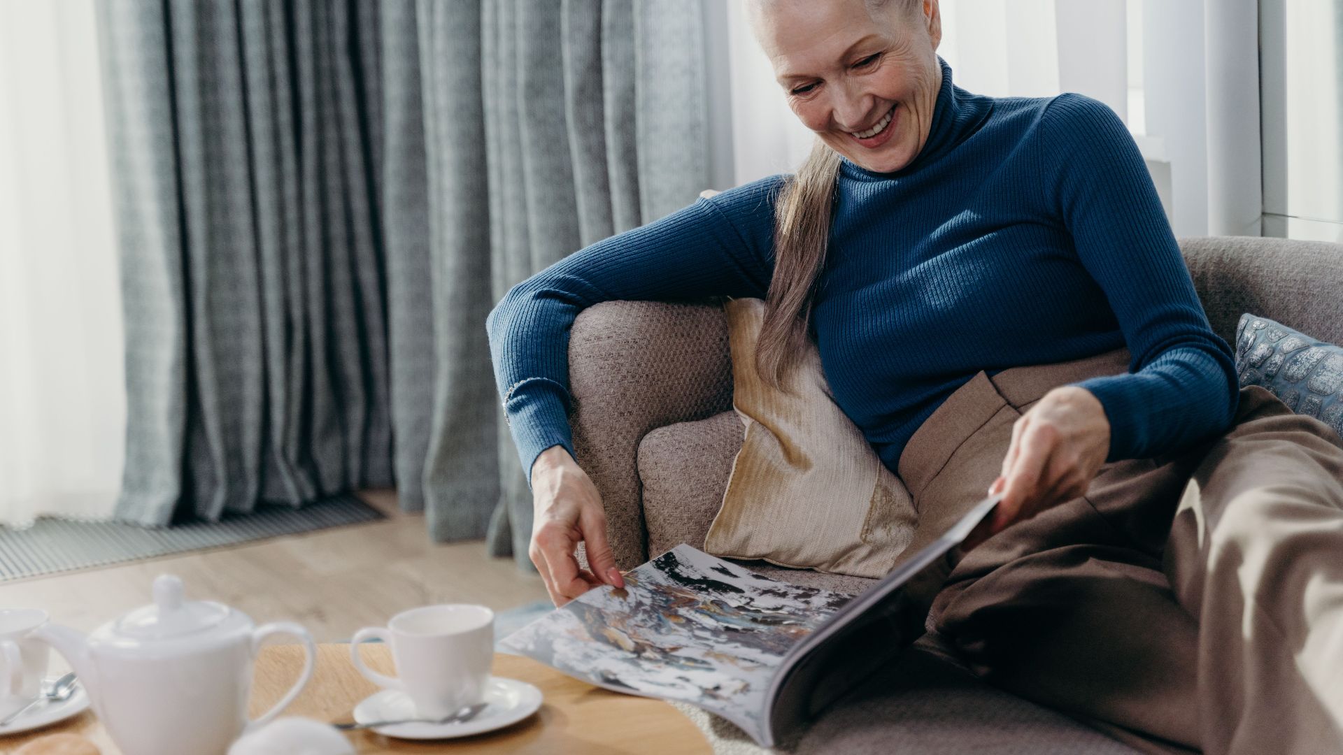 Elderly woman smiling while reading a magazine on a sofa, enjoying a cozy afternoon.