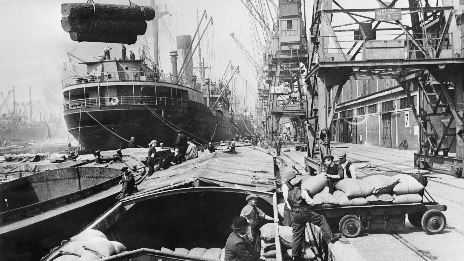 Scene in a London Dockyard during the Second World War
Dockers transfer imported and exported goods by hand, vehicle and crane between merchant ships and river barges at a London Dockyard. Comment: See wording on the crane. 'East Ferry'. This could be Mil