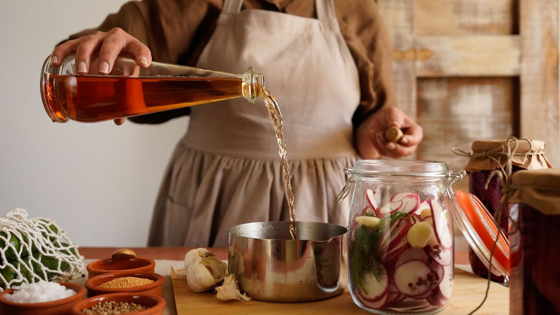 A woman pours vinegar into a pot while making homemade pickles in a rustic kitchen setting.