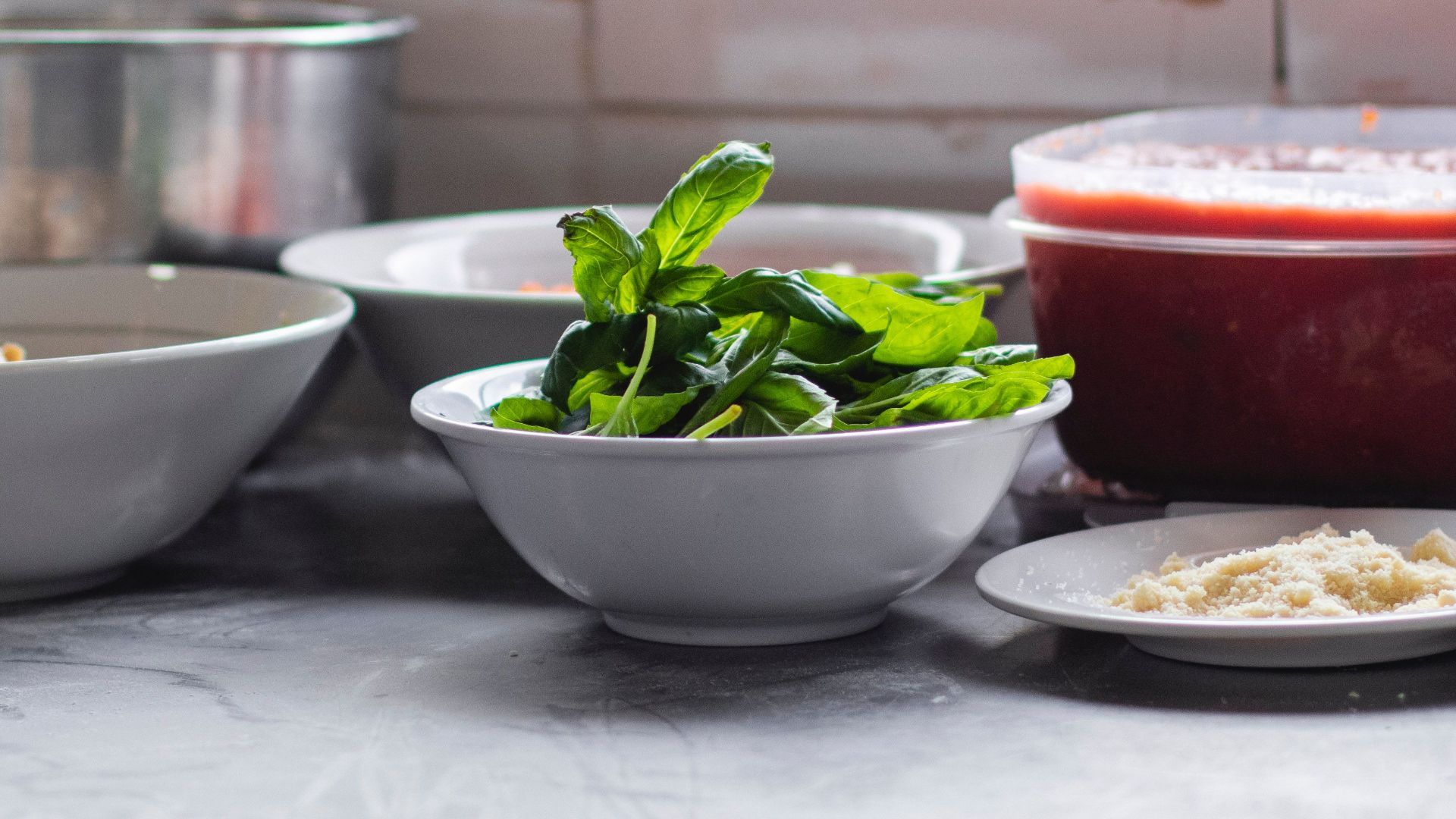 Preparation scene with fresh basil, parmesan, and tomato sauce on a kitchen counter.