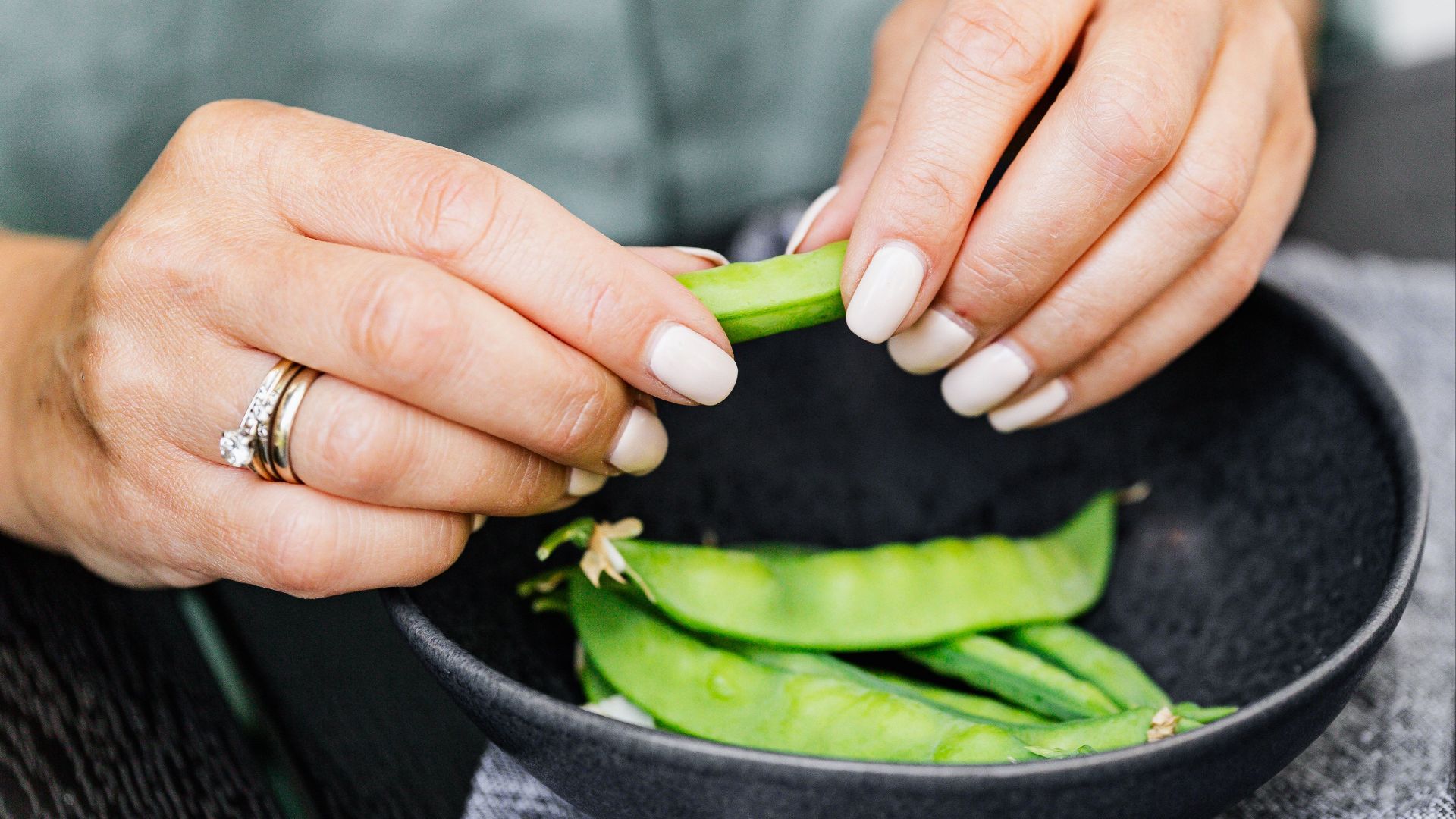A woman with manicured nails shells fresh snap peas into a black bowl.