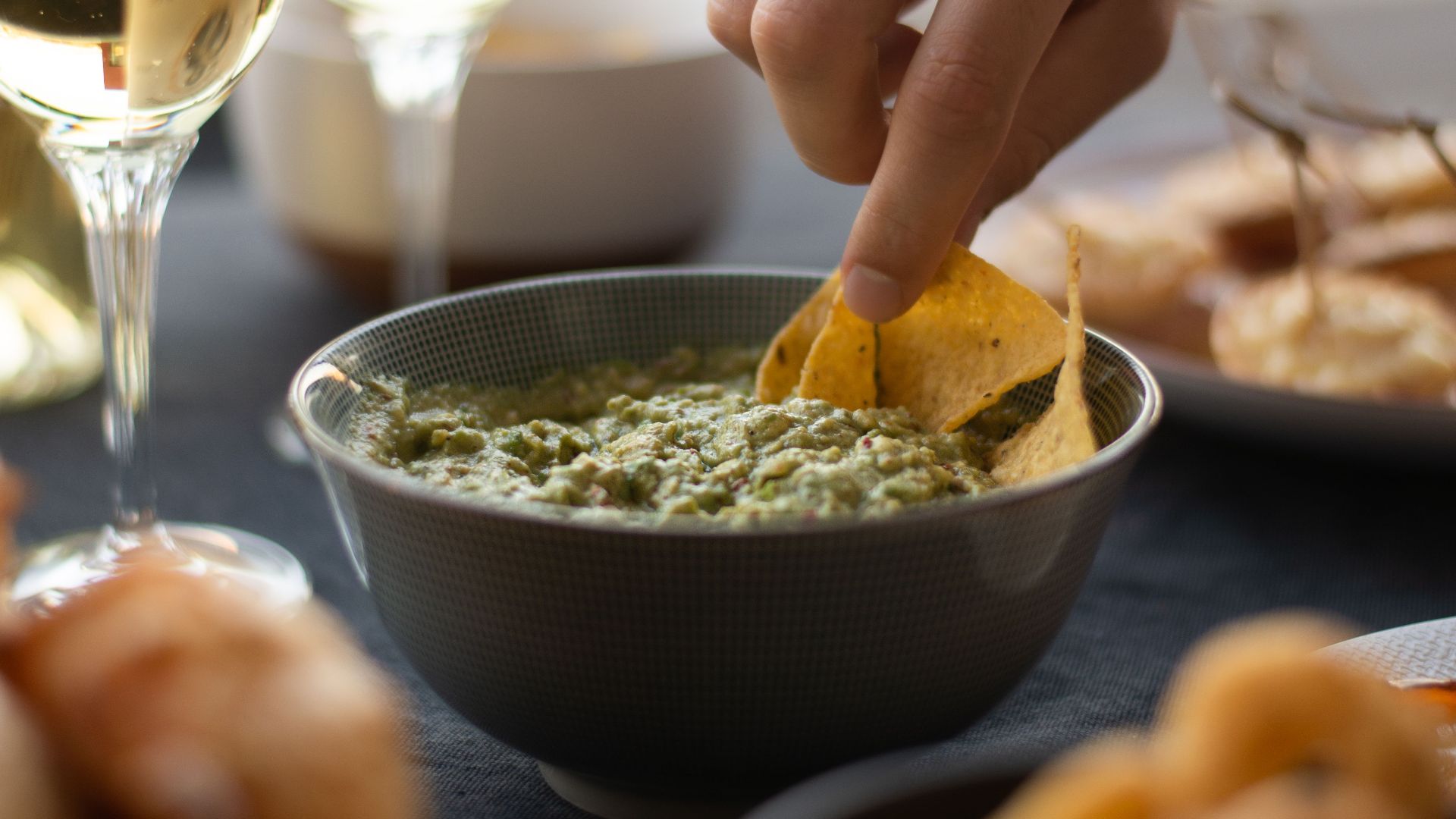 A hand dipping nachos into a bowl of fresh guacamole, accompanied by wine glasses.