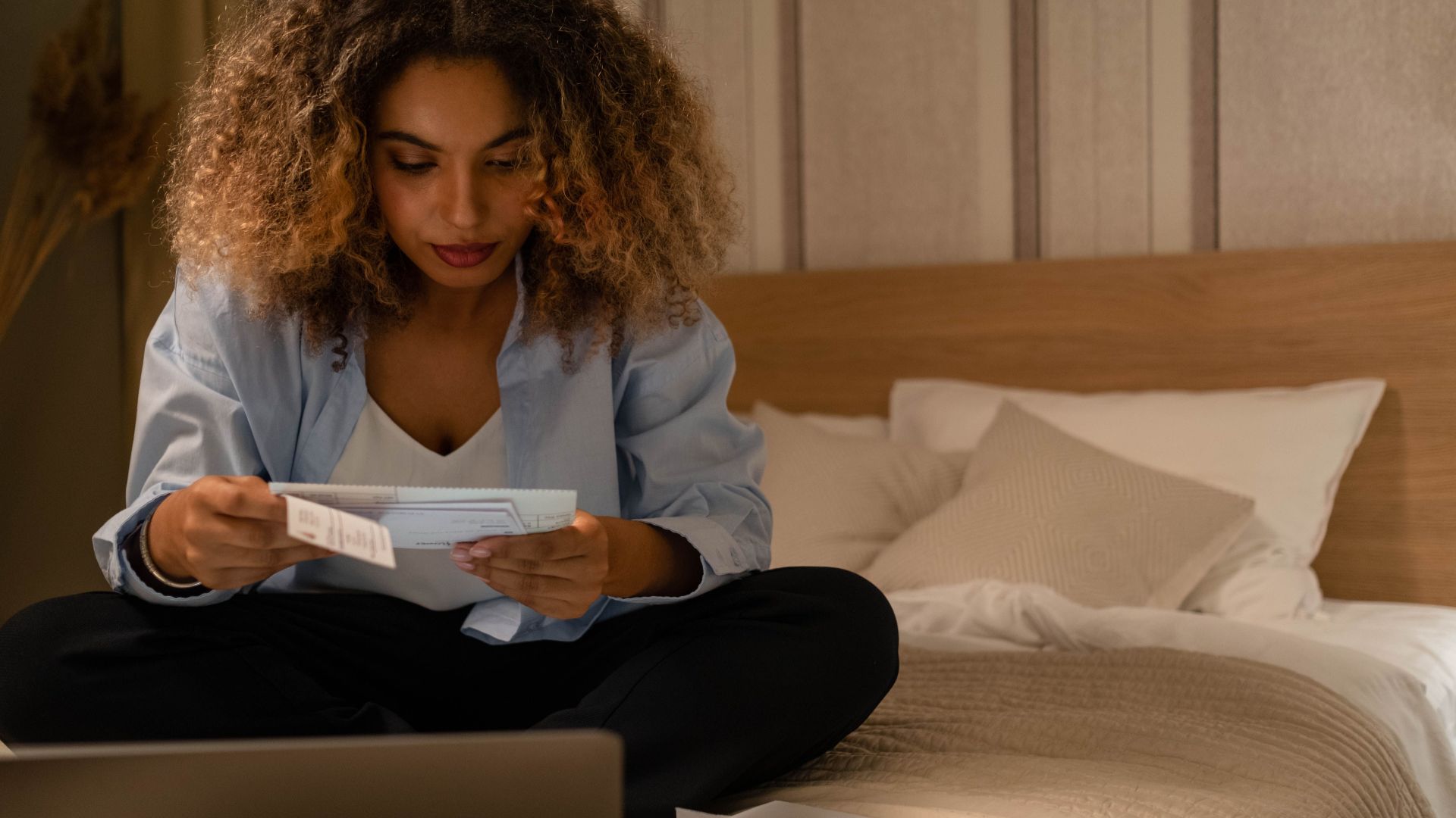 Woman sitting on bed reading documents with laptop, highlighting home office setting.